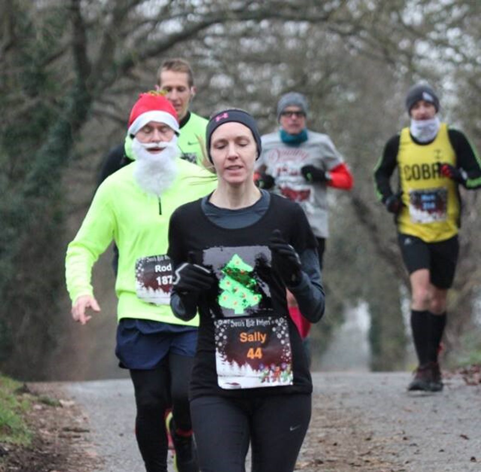 A group of runners participate in a road race. The nearest runner, "Sally," wears a black outfit with a Christmas tree design. Behind her, a runner in a Santa hat and fake beard stands out in bright yellow. Other runners are in various athletic gear.