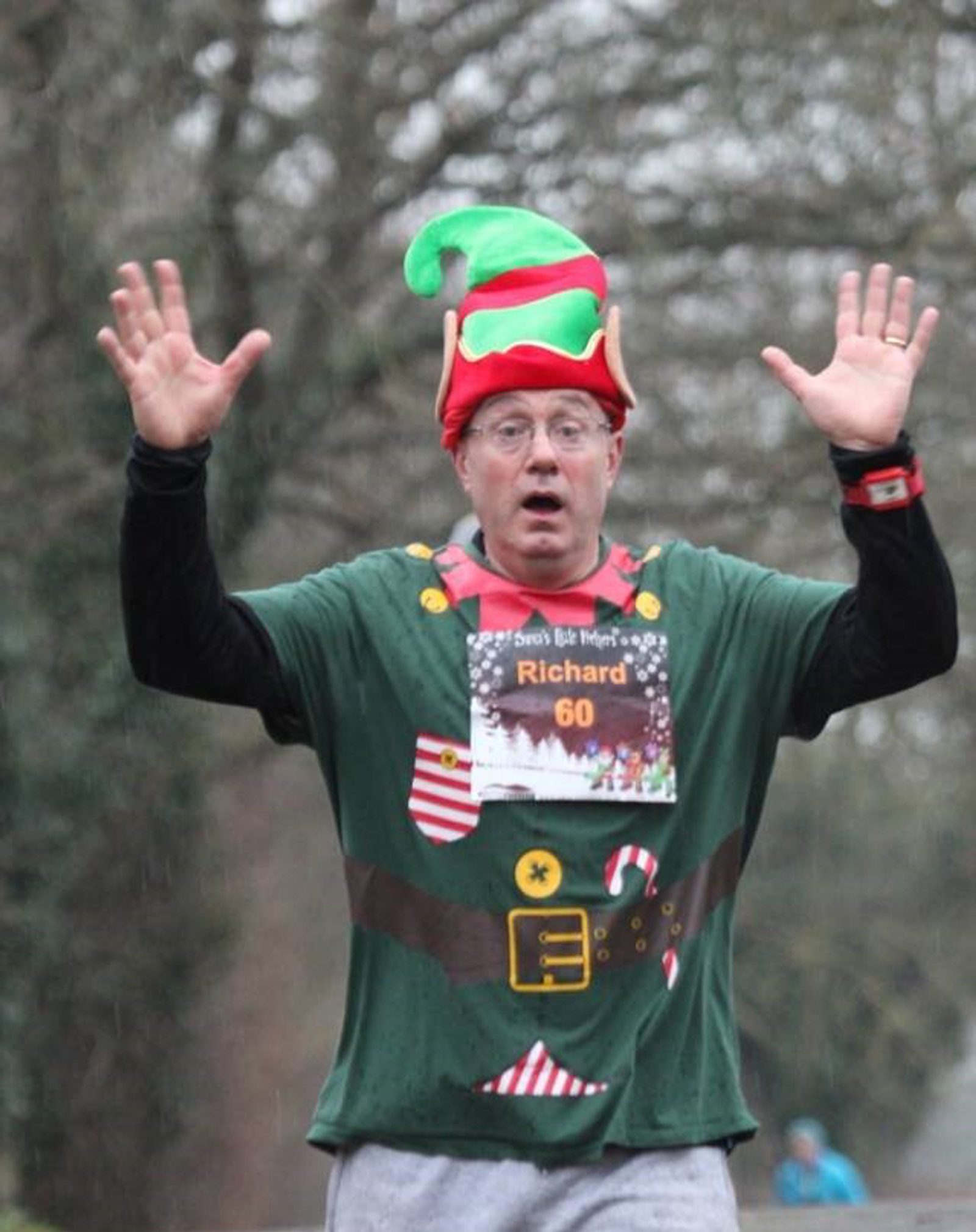 A person dressed in an elf costume with a festive hat and T-shirt participates in a race. Number 60 and the name "Richard" are displayed on their bib. They have their hands raised and are in an outdoor setting with trees in the background.