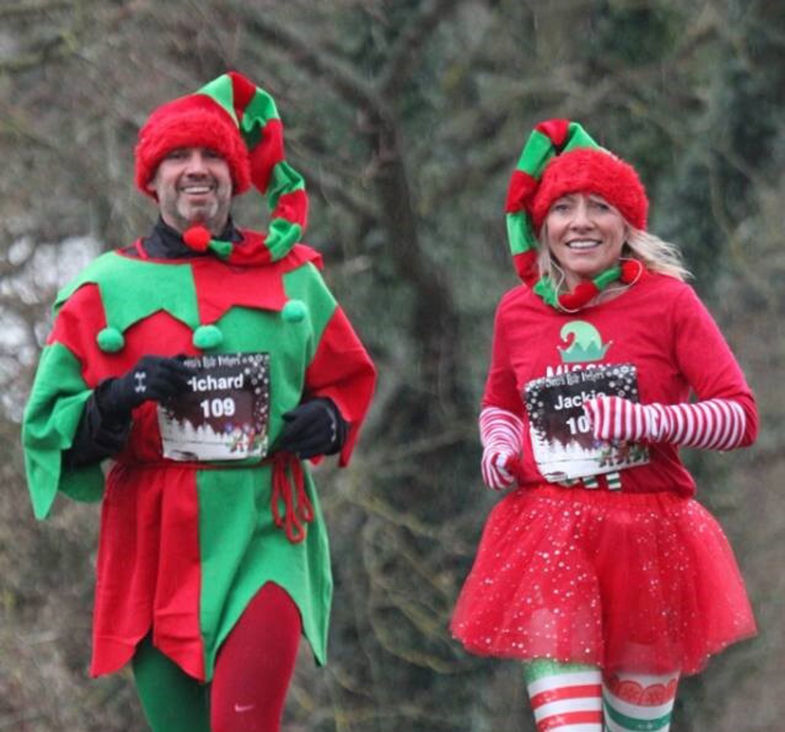 Two people jog on a path dressed in festive attire. The person on the left wears a green and red elf costume with a pointy hat, while the person on the right wears a red tutu, striped arm warmers, and a red hat. Both have race bibs on their shirts. The background is a blurred, wooded area.