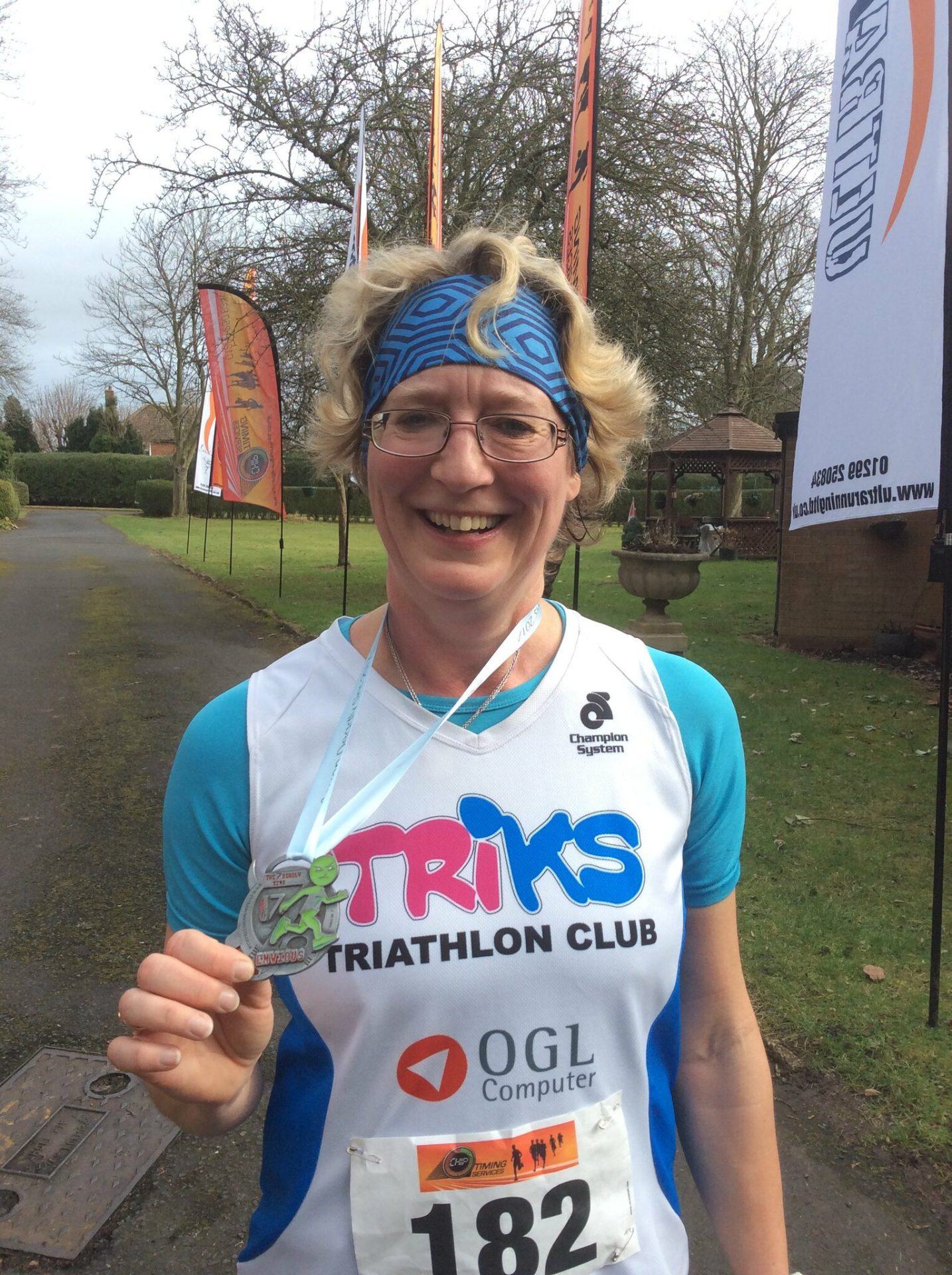 A smiling individual wearing a blue headband and a "TriKS Triathlon Club" jersey holds up a medal. They are outdoors, with colorful flags and a race number (182) visible. Trees and a grassy area can be seen in the background.