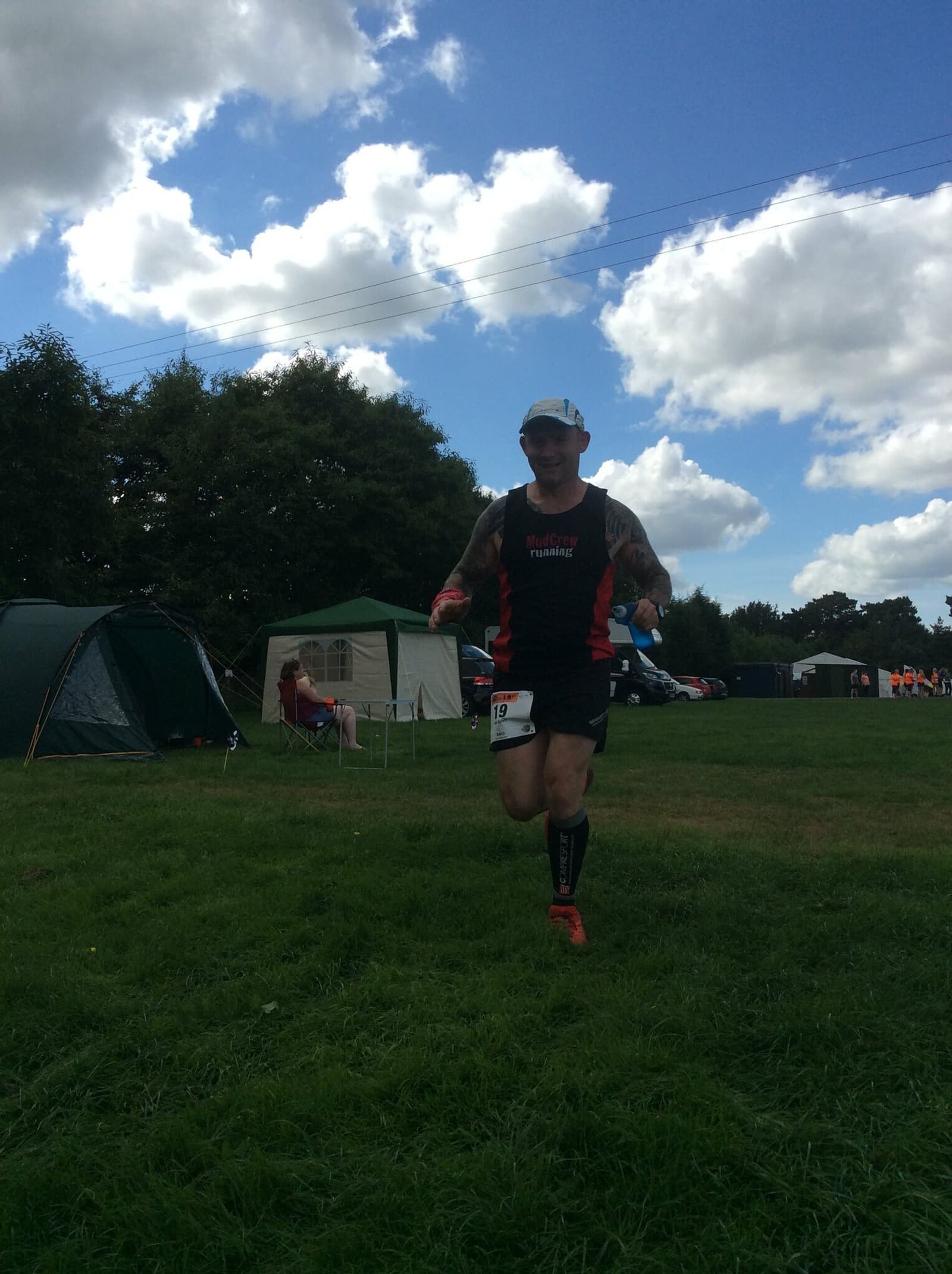 A runner wearing a black and red tank top, black shorts, and a white cap participating in a race on a grassy field. The runner has a race number 19 and is making their way past tents and spectators, with a partly cloudy sky in the background.