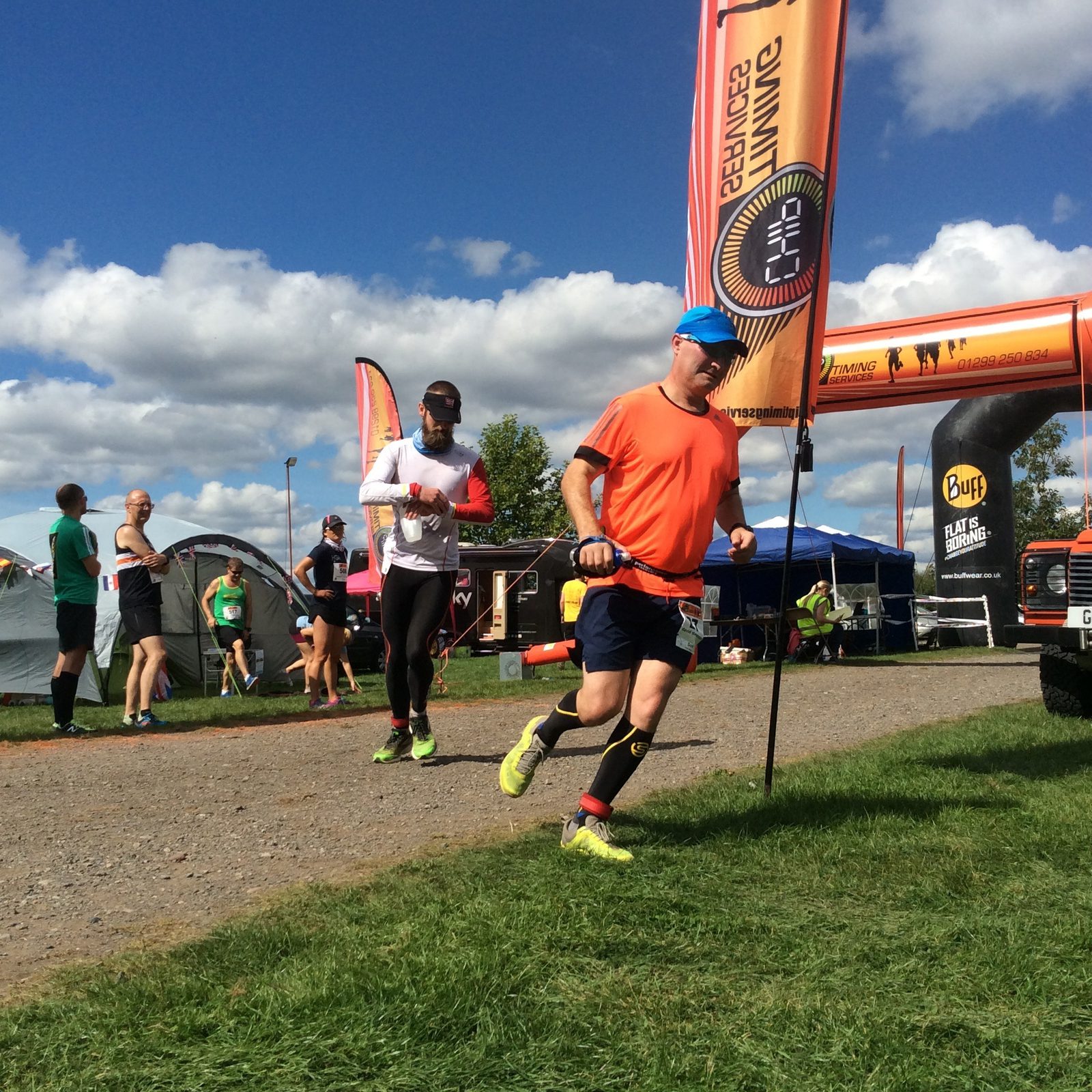 A group of runners participate in an outdoor race during a sunny day. The nearest runner, wearing an orange shirt and blue cap, is in the foreground with a focused expression. Banners and tents are set up along the grassy path, and spectators are visible in the background.