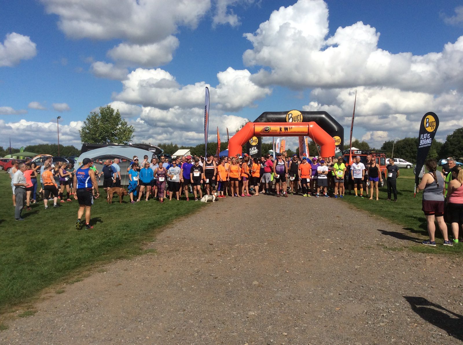 A group of runners gather at the starting line under an orange inflatable arch on a grassy field. The sky is blue with scattered clouds. Spectators, some holding flags and banners, stand on the sides. The event name, "RUN," is visible on the arch.
