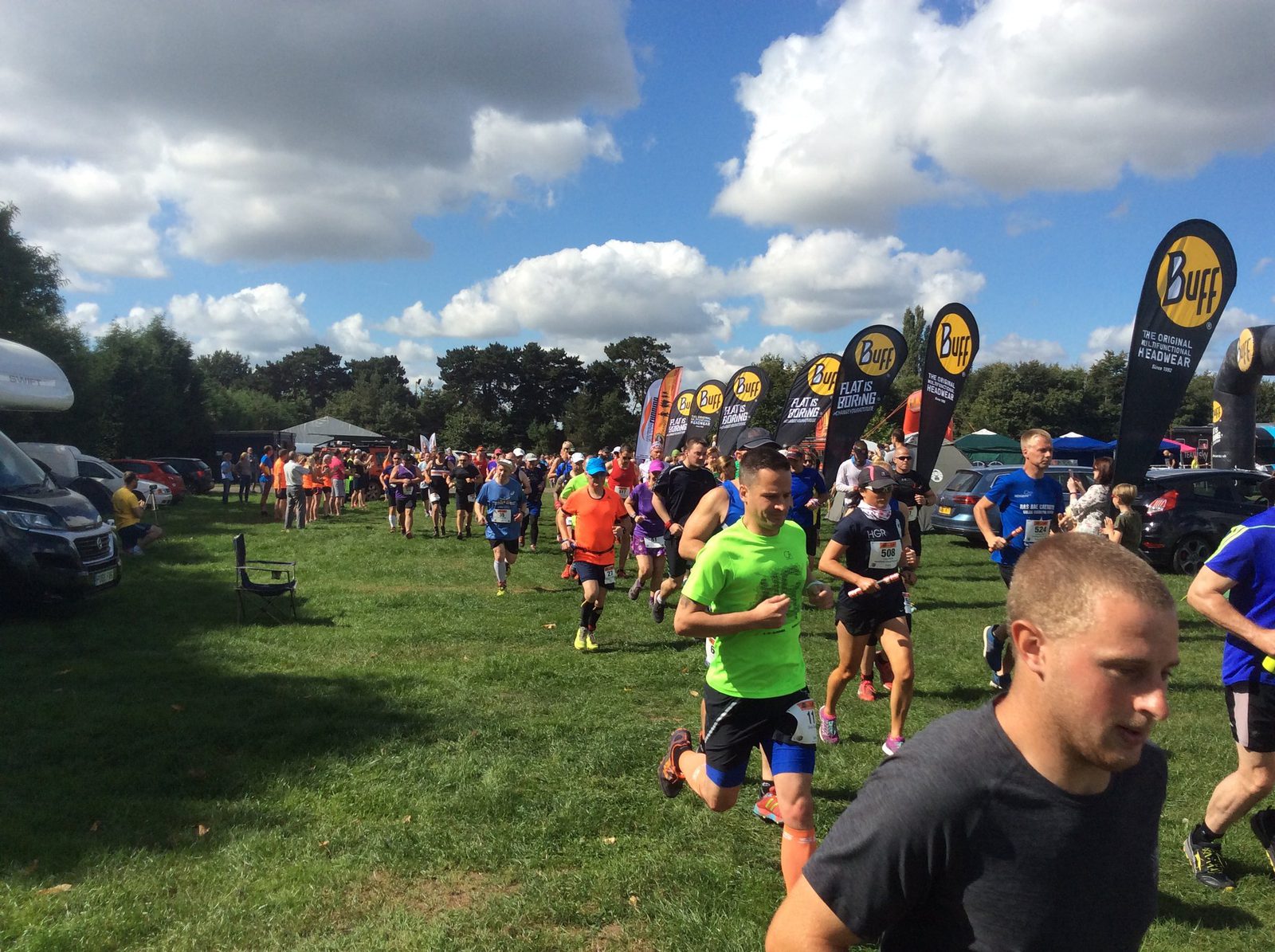 A group of runners participating in an outdoor race run across a grassy field under a partly cloudy sky. Various banners with "Buff" branding are visible along the route. Spectators and parked cars can be seen in the background.