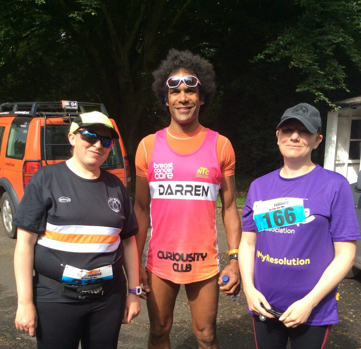 Three people are standing outdoors in athletic attire. The person in the middle has a race bib reading "DARREN" and is wearing a pink outfit. The other two people are also wearing race bibs and sunglasses. There is a vehicle and trees in the background.