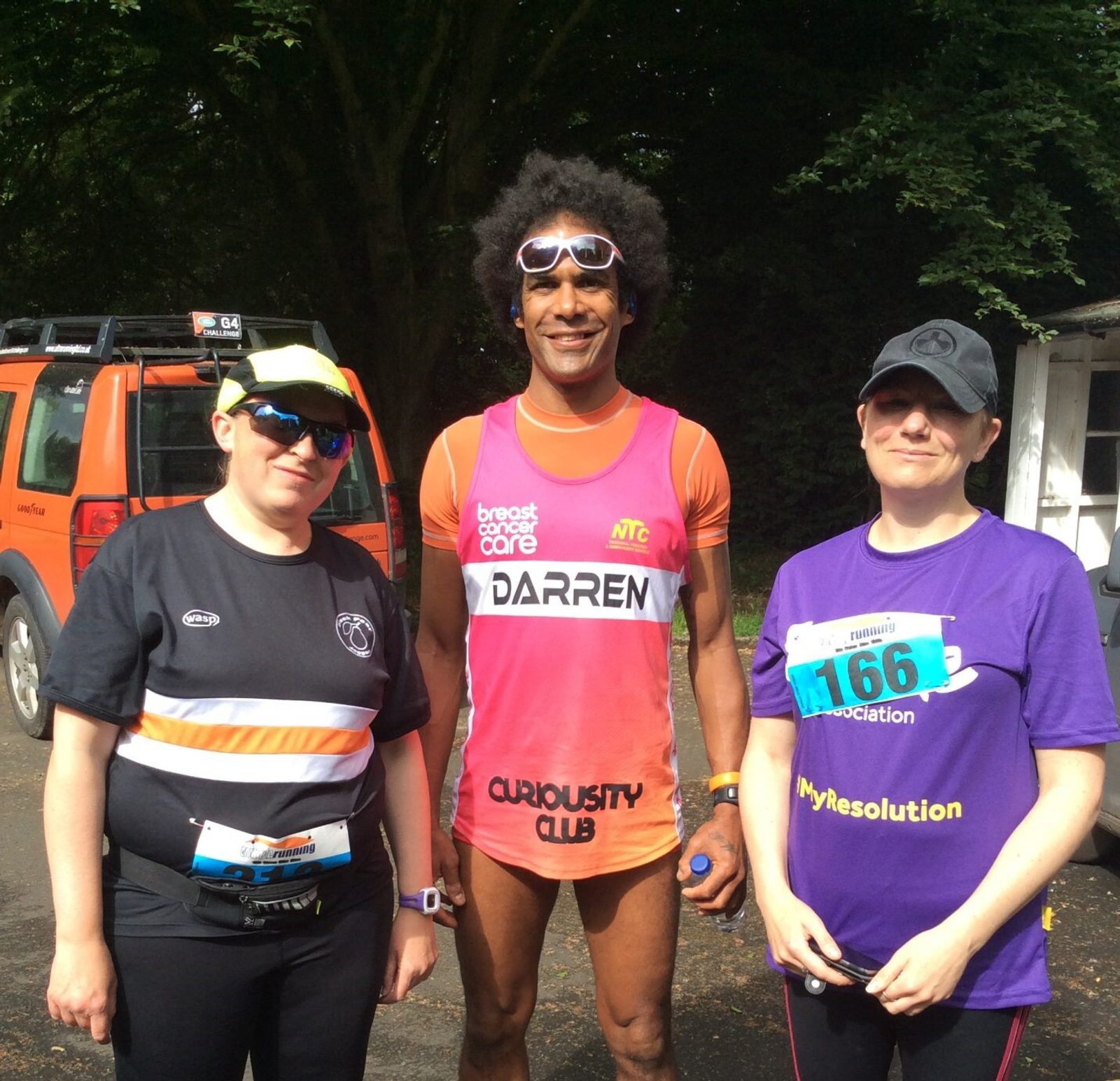 Three people are standing outdoors in athletic attire. The person in the middle has a race bib reading "DARREN" and is wearing a pink outfit. The other two people are also wearing race bibs and sunglasses. There is a vehicle and trees in the background.