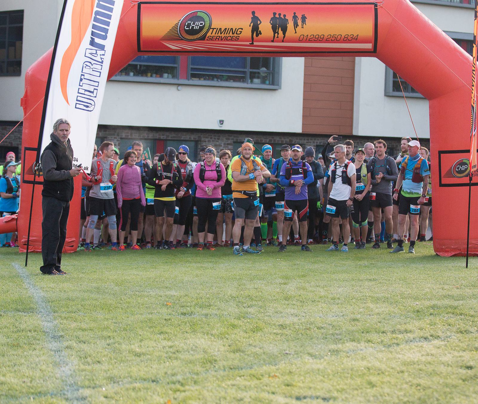 A group of runners stand at the starting line under an inflatable arch with banners that read "Ultra Running" and a banner displaying "Chip Timing Services" with a phone number. The runners are in athletic gear, and a person stands to the side, possibly giving directions.
