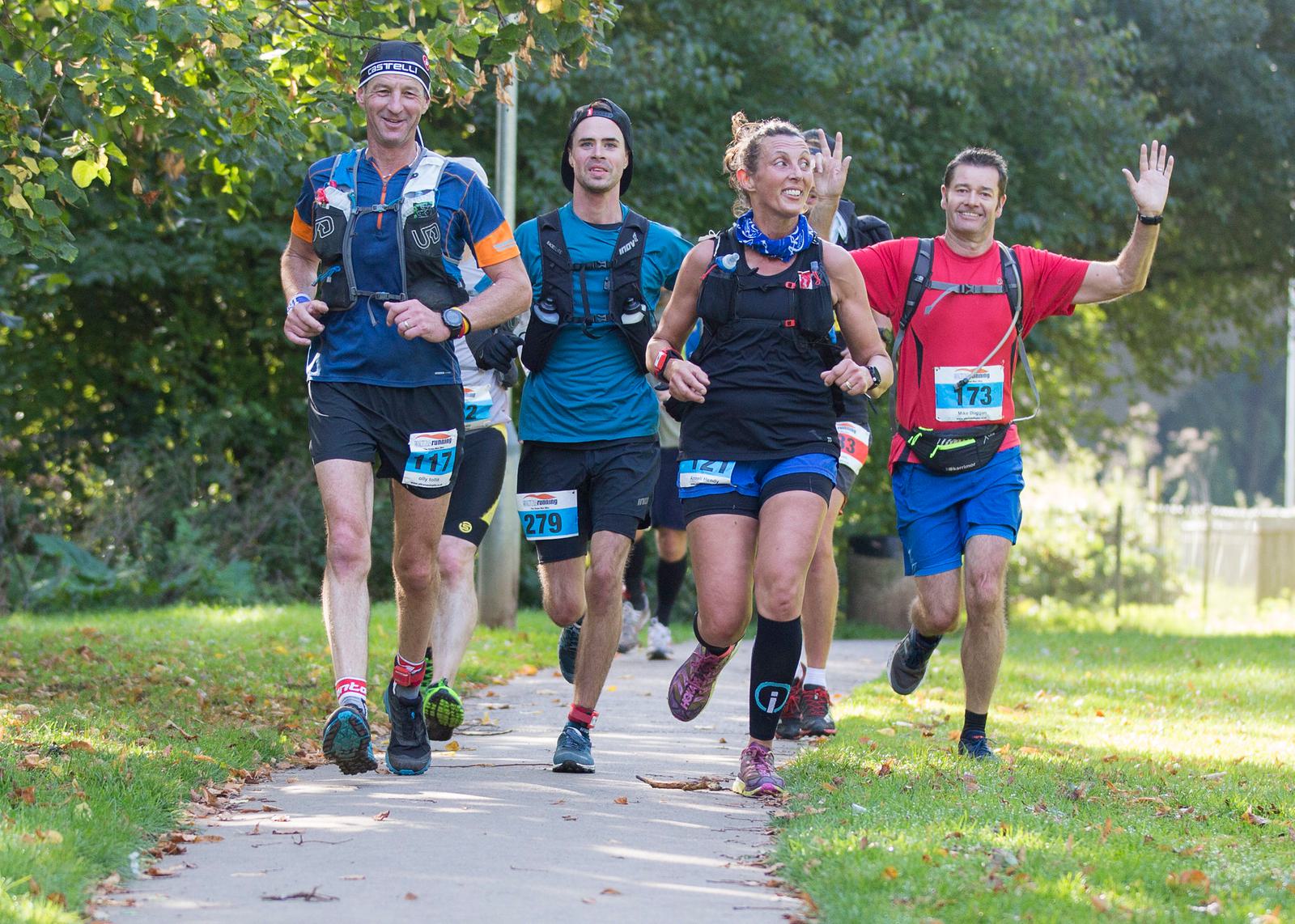 Four runners are on a sunny park trail during a race. Two men and two women wear race bibs and running gear. One man waves at the camera. The group appears cheerful and energetic as they run amongst green trees and grass.