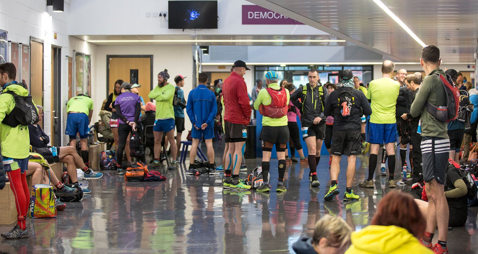 A group of people, mostly dressed in athletic attire such as shorts, shirts, and running shoes, gather in a brightly lit indoor space. Some are standing, others sitting and organizing gear. It appears to be the start or end of a sporting event.
