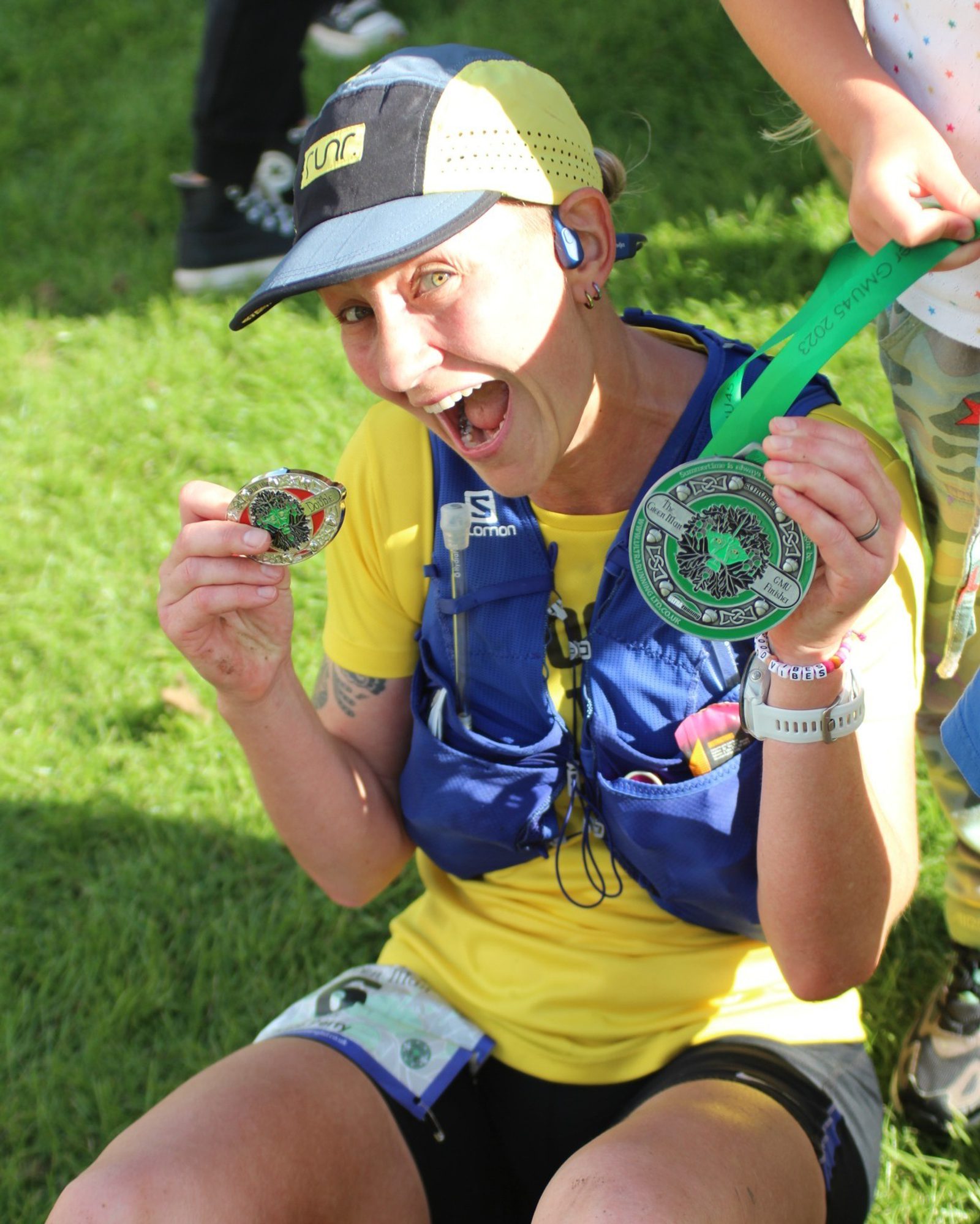A person in a yellow shirt and blue vest, wearing a cap, smiles widely while holding two medals. They are seated on grass, with another person partially visible beside them.