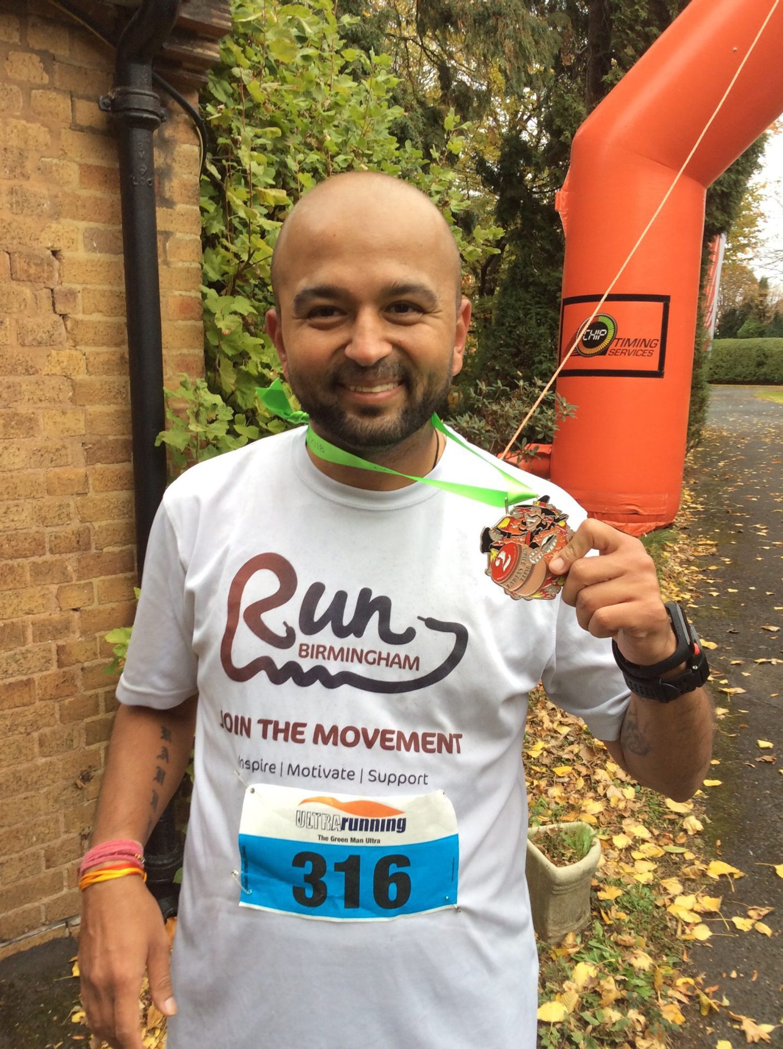A cheerful man with a shaved head and beard proudly holds up a medal after finishing a race. He wears a white "Run Birmingham" t-shirt, a race bib numbered 316, and colorful wristbands. An orange inflatable finish line arch is visible in the background.