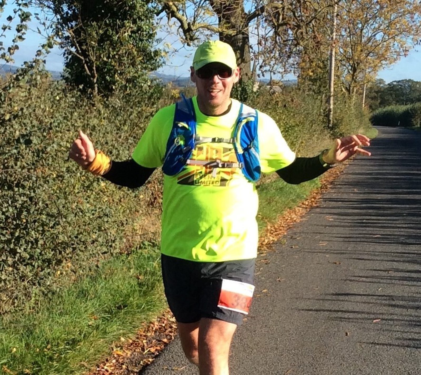 A person wearing a neon yellow shirt, cap, and a blue hydration pack is running on a rural road. The person is smiling and has their arms raised in a cheerful manner. The road is bordered by trees and bushes with autumn foliage. The weather is sunny.