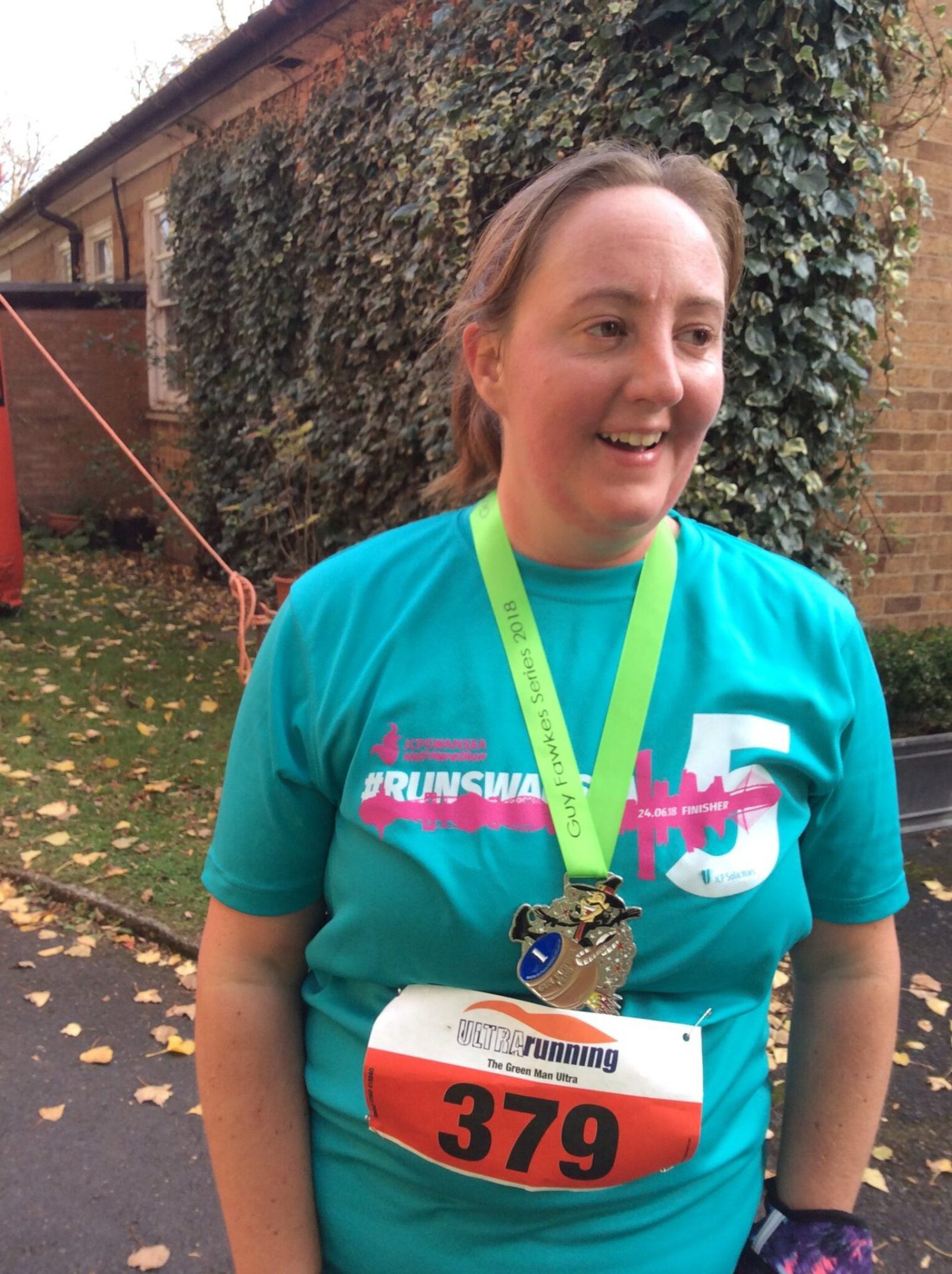 A person wearing a turquoise shirt with a "Run Sweat Love" design and an orange race bib numbered 379 smiles while displaying a gold medal on a green ribbon. They are standing outside near a building and a leafy bush, appearing to be at a race event.