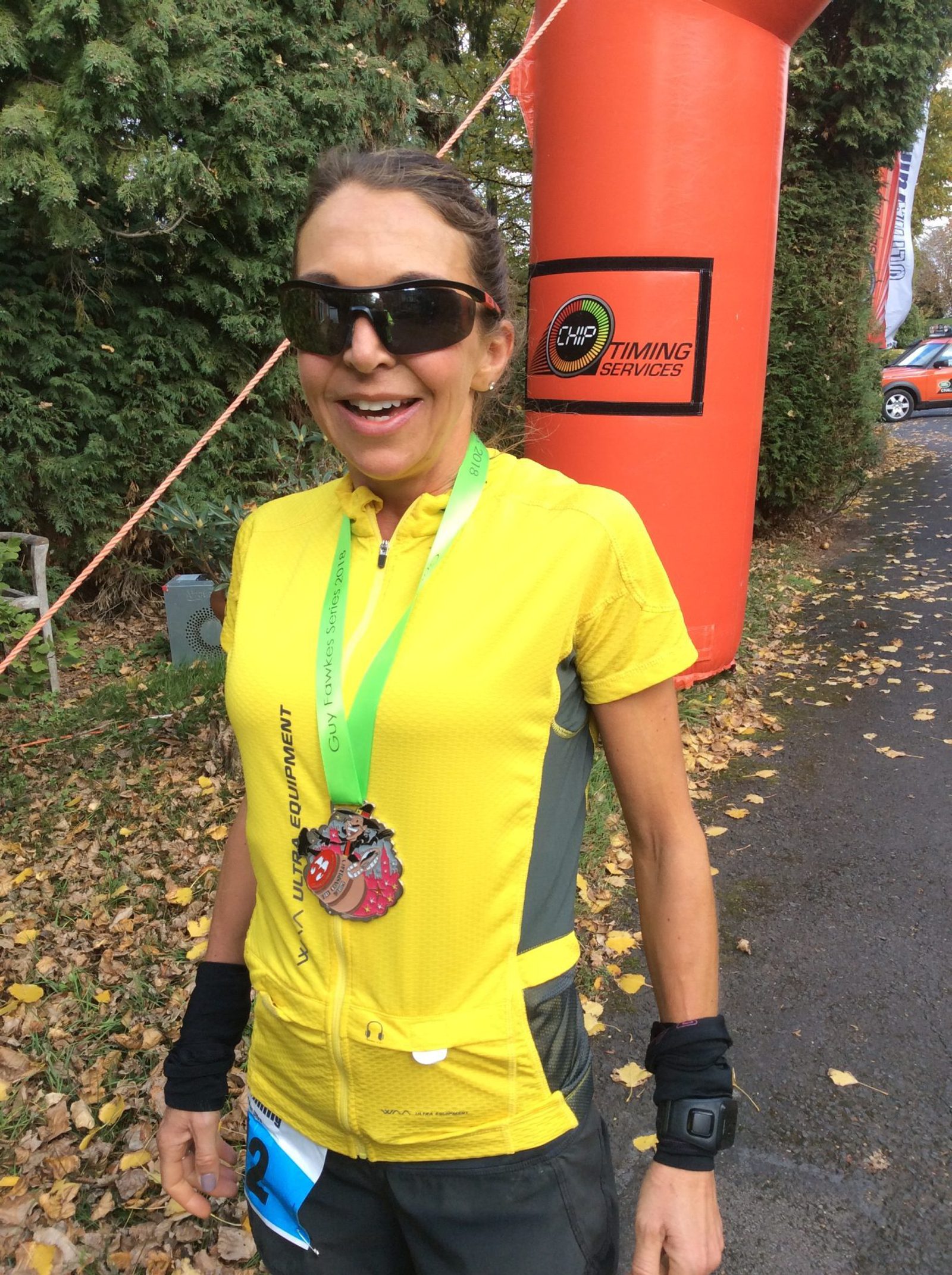 A woman wearing sunglasses and a yellow cycling shirt stands near an orange pillar outdoors. She has two medals around her neck and is smiling. The ground is covered in fallen leaves, and there is greenery in the background along with a small sign for “Chip Timing Services.”.