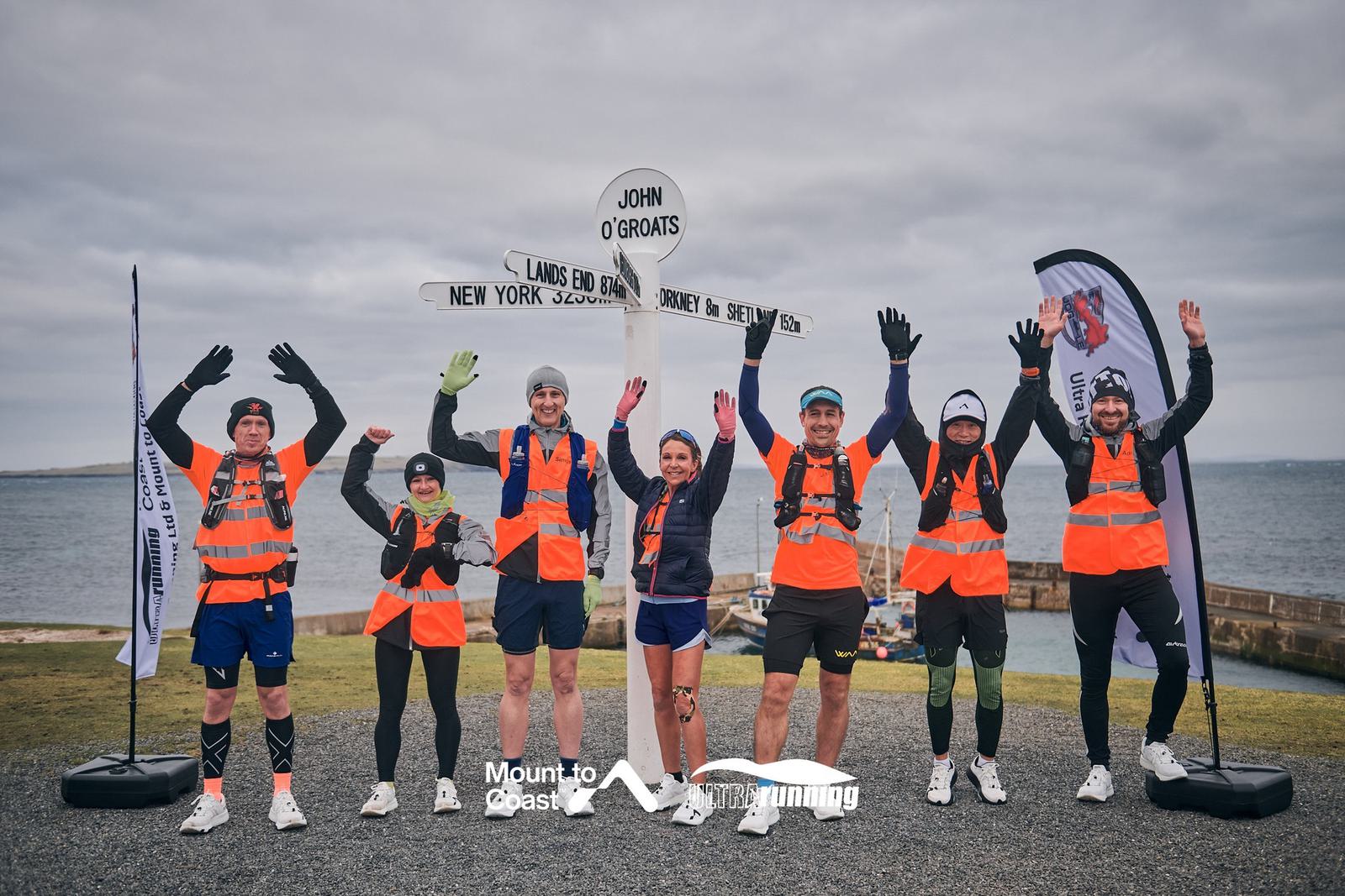Seven people wearing orange vests celebrate at John O'Groats, standing in front of a signpost with arms raised. The background includes a cloudy sky and coastline. A flag promoting an event is visible on the right.