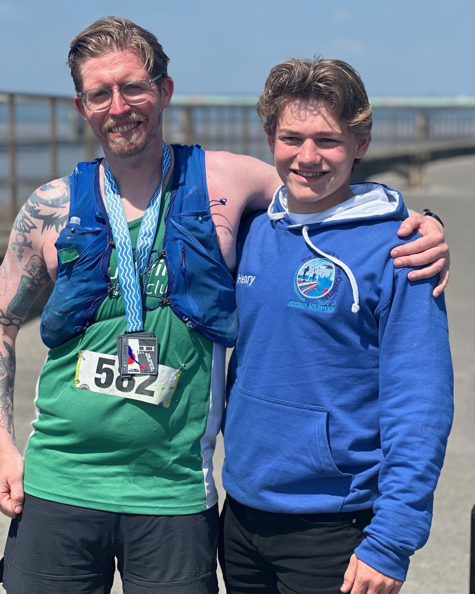 Two men, smiling. The man on the left wears a green tank top, numbered 582, with a medal and a blue hydration pack. The man on the right wears a blue hoodie with "Harvey" embroidered. They stand outdoors on a sunny day.