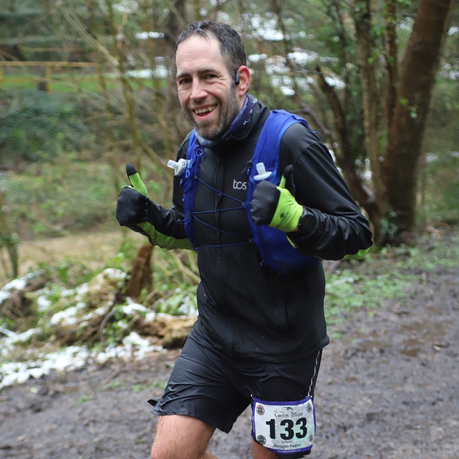 A man running in a race through a muddy, wooded trail. He's wearing a blue hydration vest, black athletic clothing, and green gloves. He smiles and gives a thumbs-up with both hands. His race number, 133, is attached to his shorts.