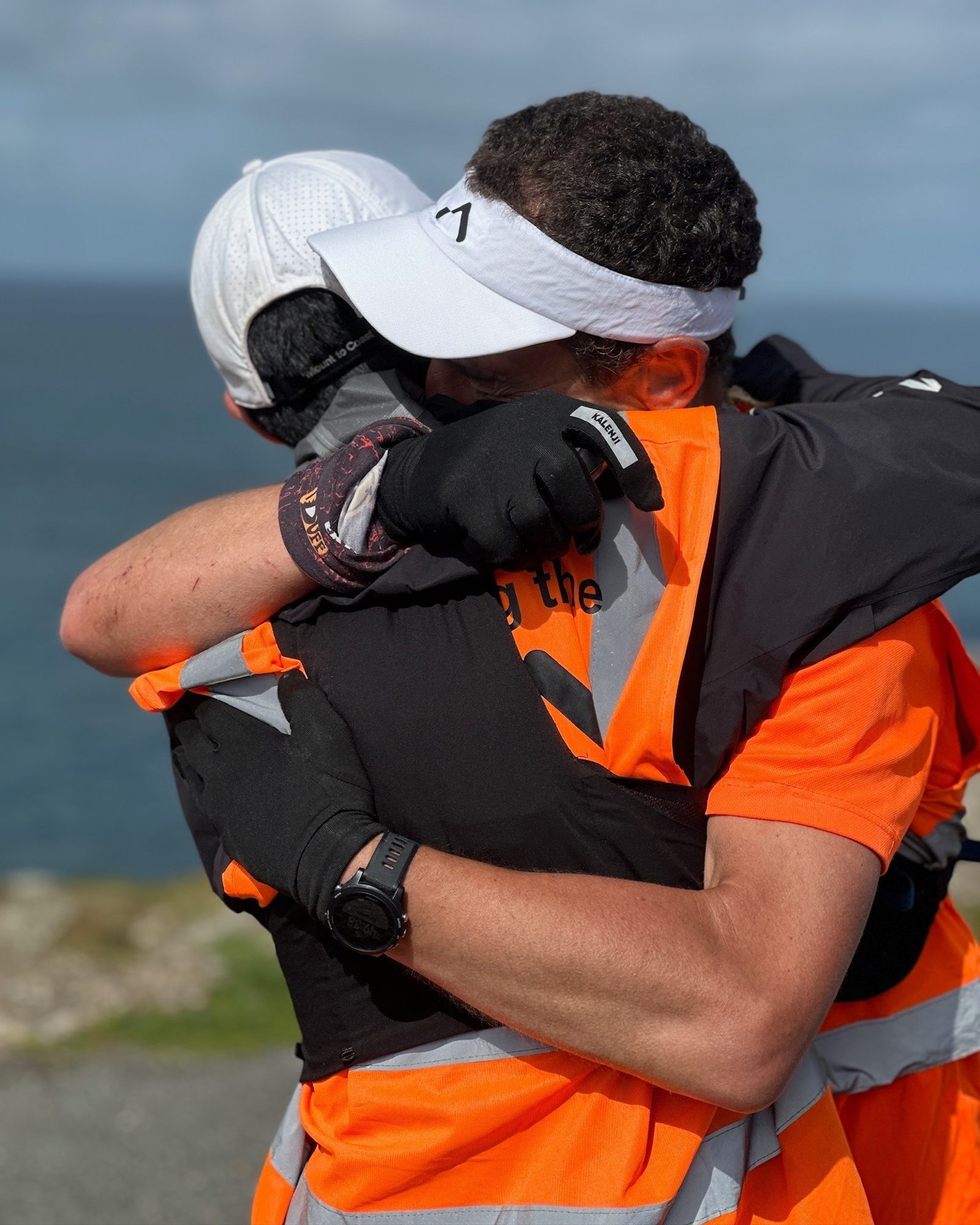 Two people in orange high-visibility gear and gloves embrace warmly, both wearing caps. They stand near a body of water under a cloudy sky.