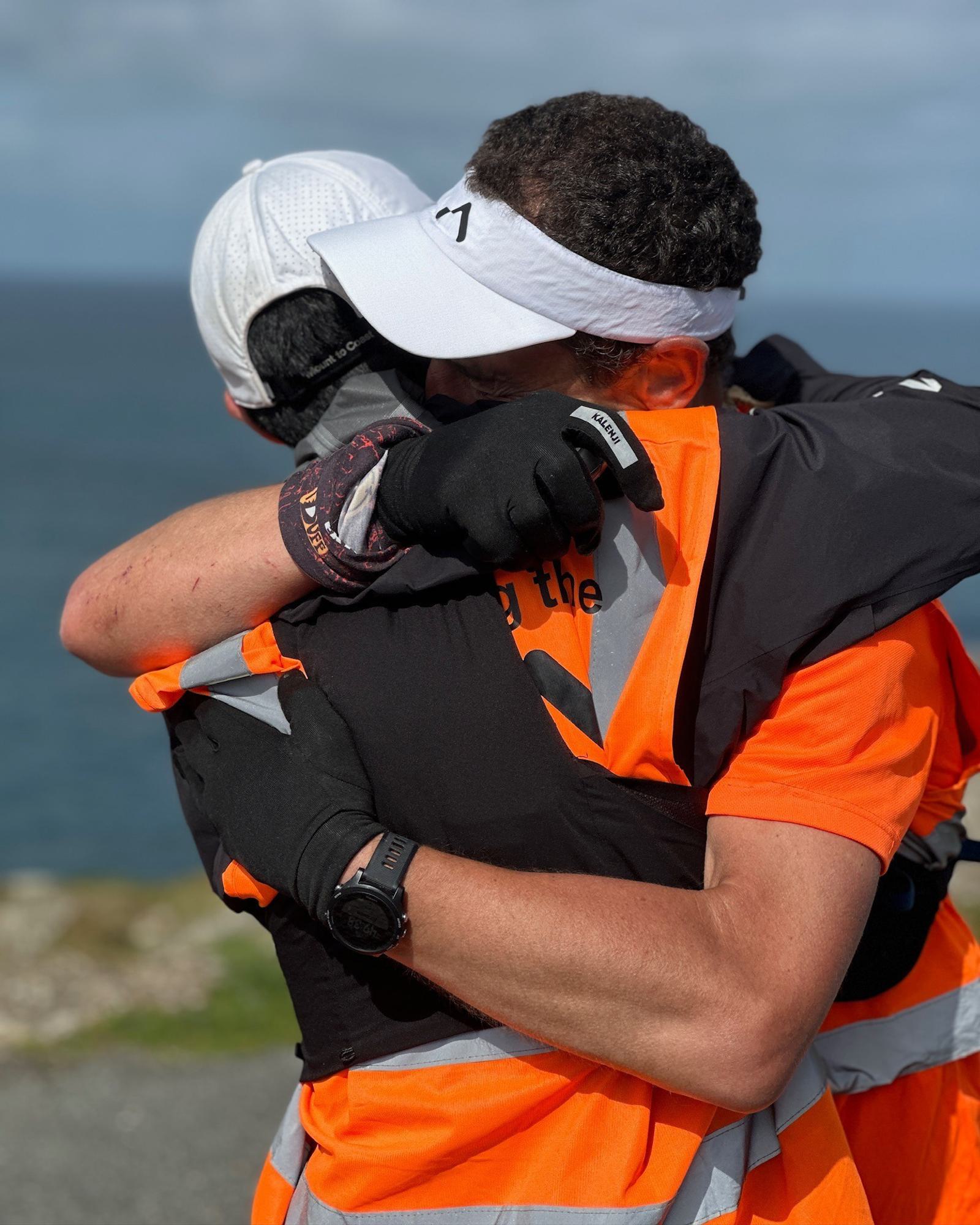 Two people in orange safety vests and athletic gear embrace warmly outdoors. One wears a white cap, the other a white visor. They stand near a coastal area under a blue sky.
