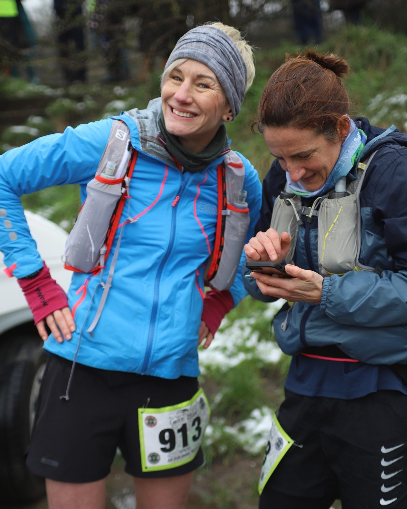 Two women in athletic gear with numbered race bibs stand outdoors. One is smiling with hands on hips, while the other looks at a phone. Both wear hydration packs and are dressed for cold weather. Snow is visible on the ground in the background.