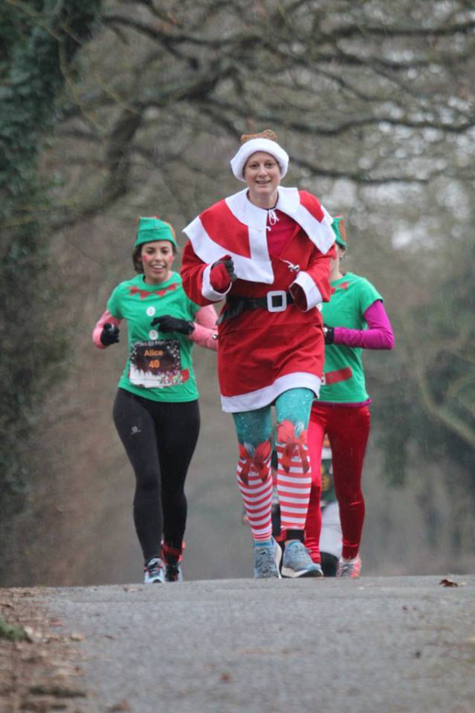 Three people are running outdoors. The person in front is dressed as Santa Claus, and the two behind are wearing green elf costumes. The background features trees and a paved path, suggesting a festive-themed race or fun run.