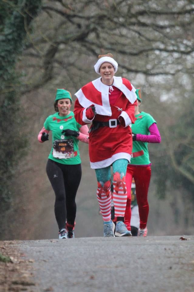 Three people are running outdoors. The person in front is dressed as Santa Claus, and the two behind are wearing green elf costumes. The background features trees and a paved path, suggesting a festive-themed race or fun run.
