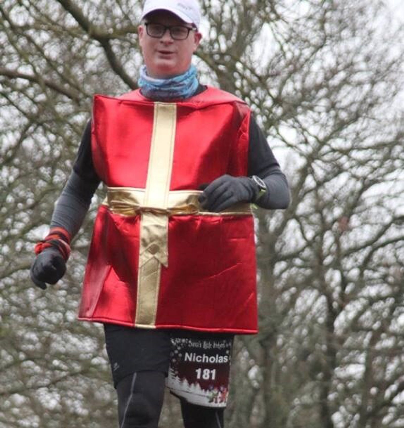 A person running outdoors, dressed in a red costume resembling a wrapped gift with a gold ribbon. They are wearing a white cap, glasses, gloves, and have a race bib pinned to their shorts with the visible number 181. The background consists of leafless trees.
