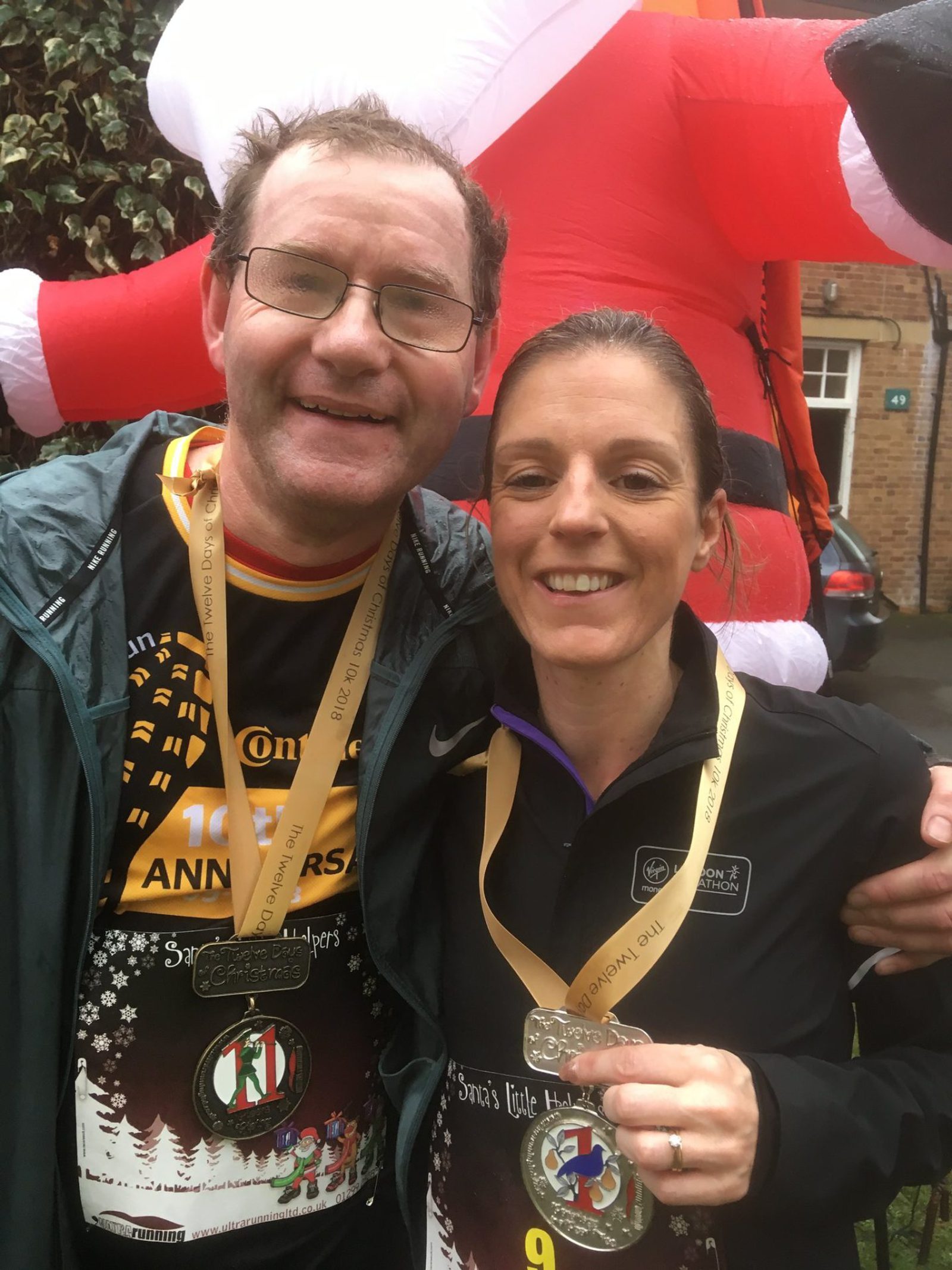 A man and a woman, smiling, pose with medals around their necks. They both are wearing event bibs and athletic clothing. An inflatable Santa Claus decoration is visible in the background, along with some greenery and parts of a brick building.
