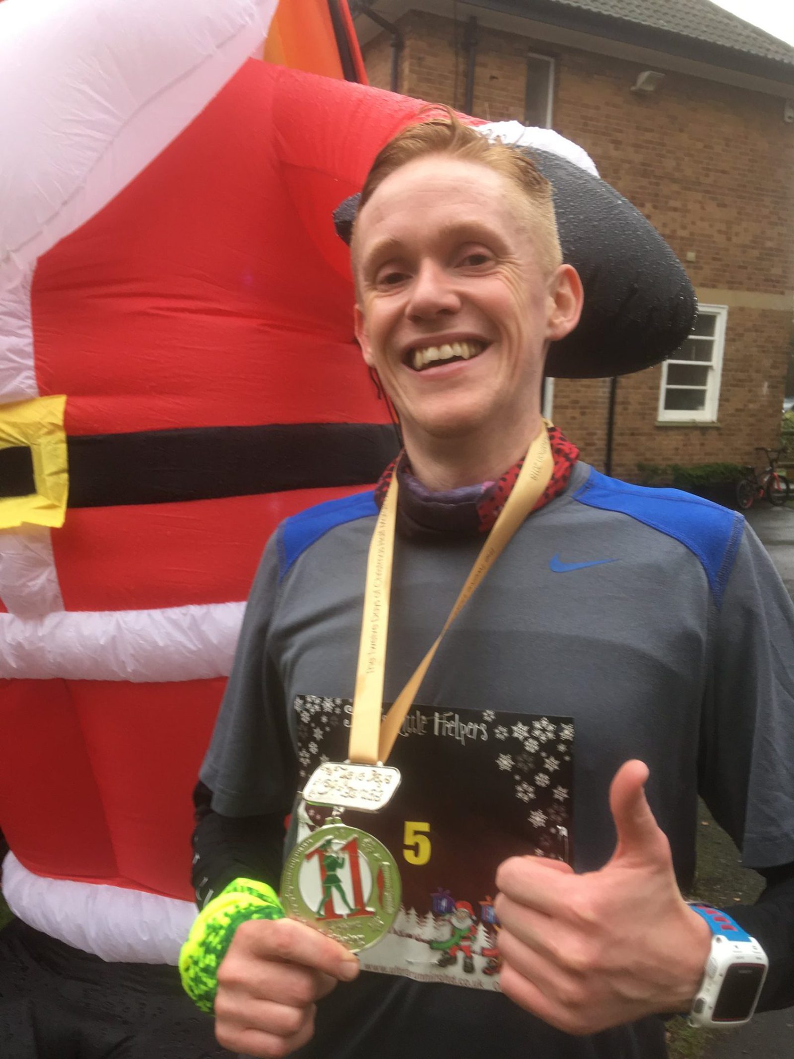 A smiling person with short, light-colored hair is holding a medal and giving a thumbs-up. They are wearing a gray and blue athletic shirt, a wristband, and a red neck gaiter. In the background, there is a large inflatable Santa Claus.