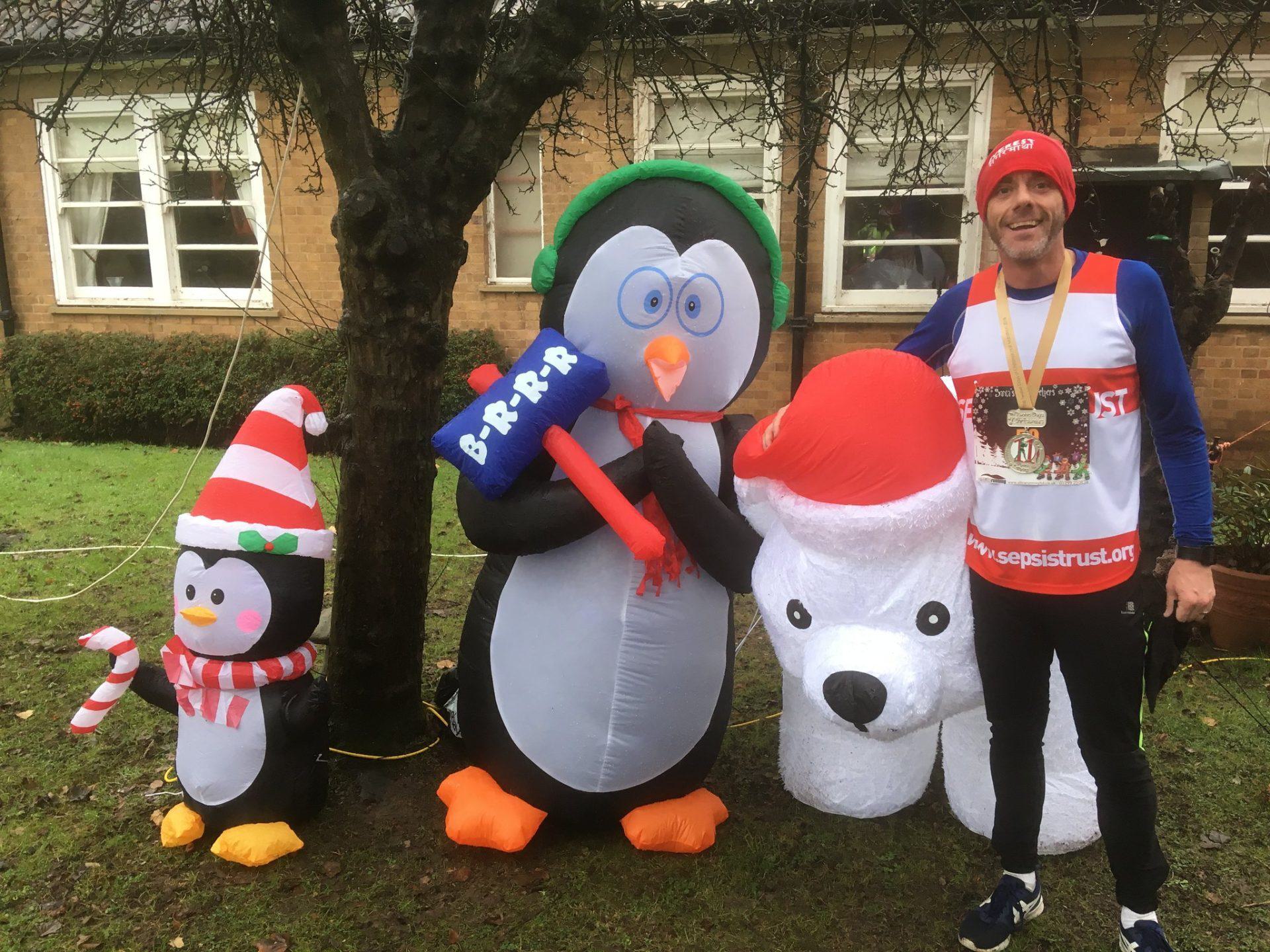 A person in running gear and a beanie stands smiling next to large inflatable Christmas decorations, including two penguins and a polar bear. The person is holding one of the polar bear's paws and is wearing a medal around their neck. The background includes a building and trees.