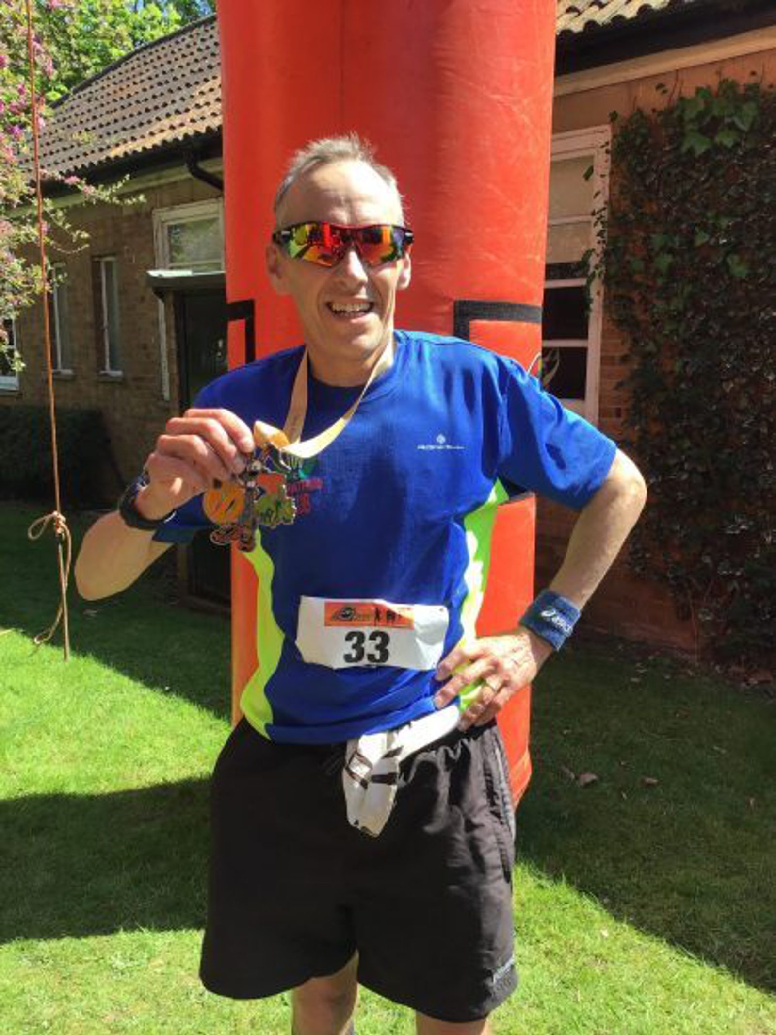 A man wearing sunglasses and a blue running shirt with neon green accents holds a medal, smiling. He has a race bib with the number 33 and stands in front of an orange inflatable column, with a house and greenery in the background.