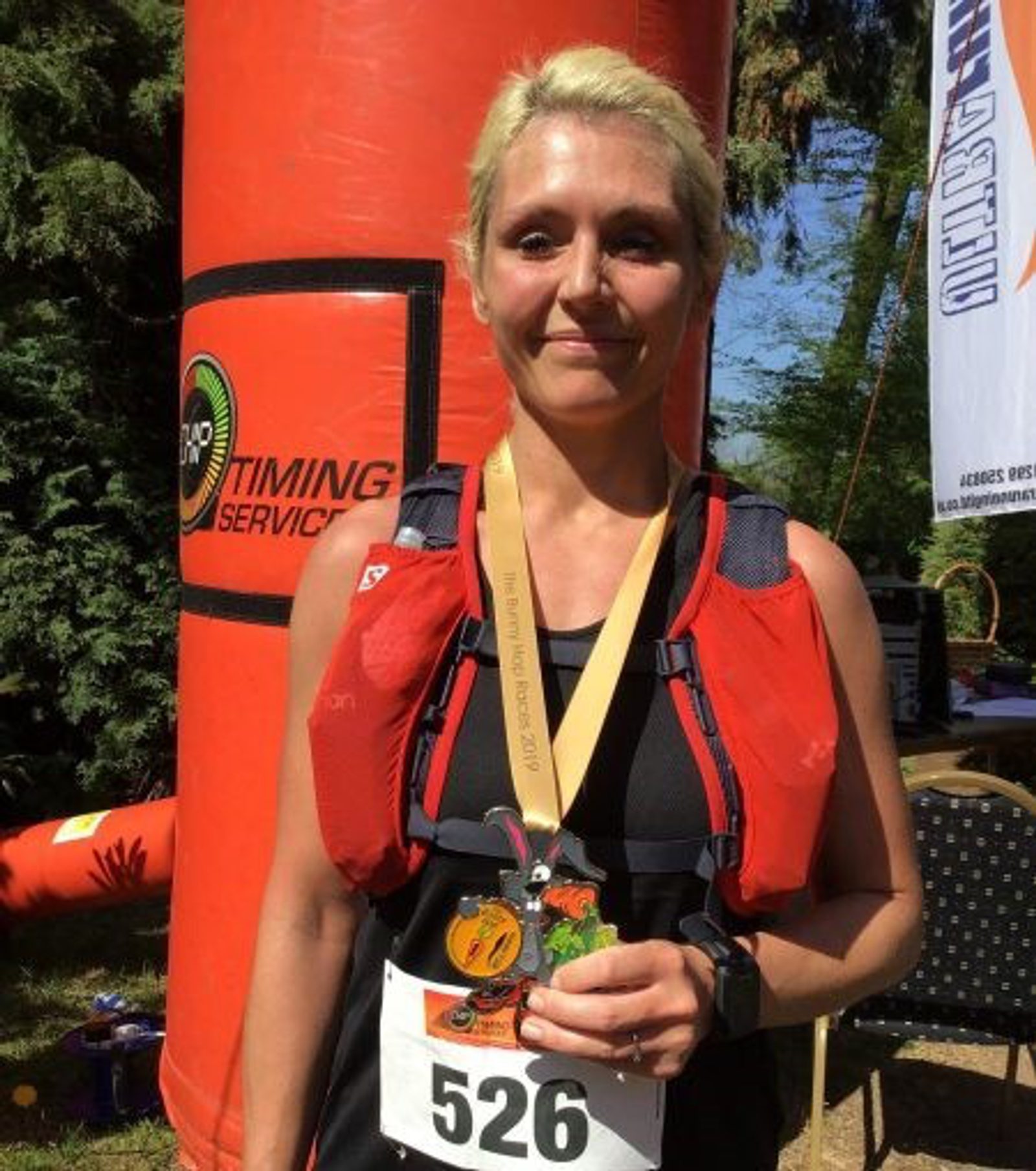 A woman with blonde hair, wearing a black outfit and red hydration pack, stands outdoors holding a medal. She has a race bib with the number 526. Behind her is an orange inflatable arch and various banners. She appears to have just completed a race.