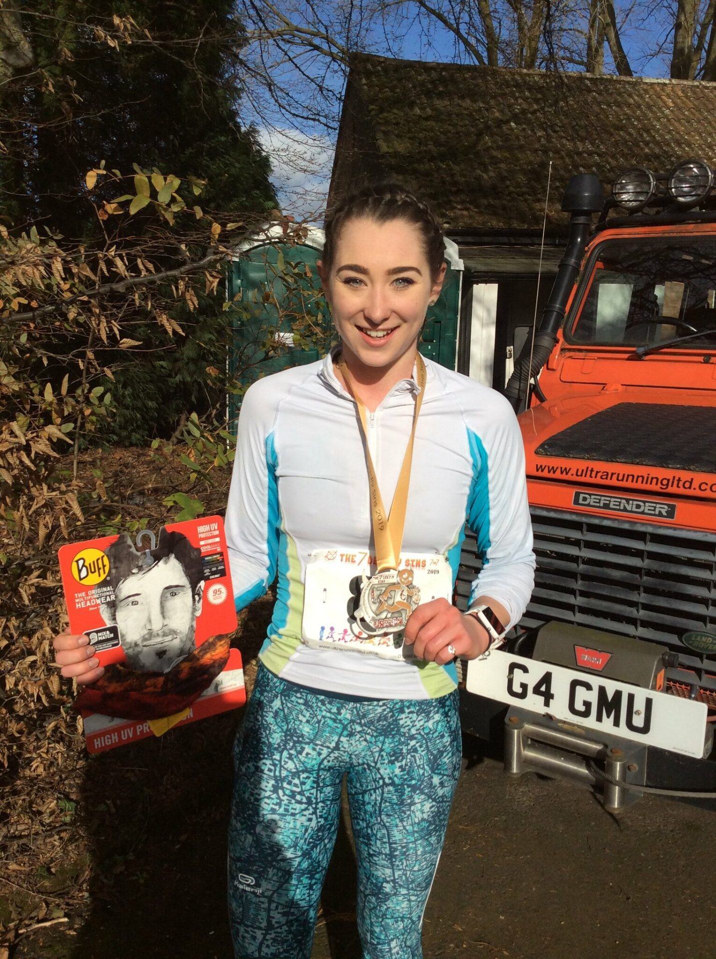A woman stands outdoors holding a running magazine and wearing a medal around her neck. She is dressed in athletic gear including a white running jacket and patterned blue leggings. Behind her is an orange vehicle with the website "ultrarunningltd.com" on the windshield.