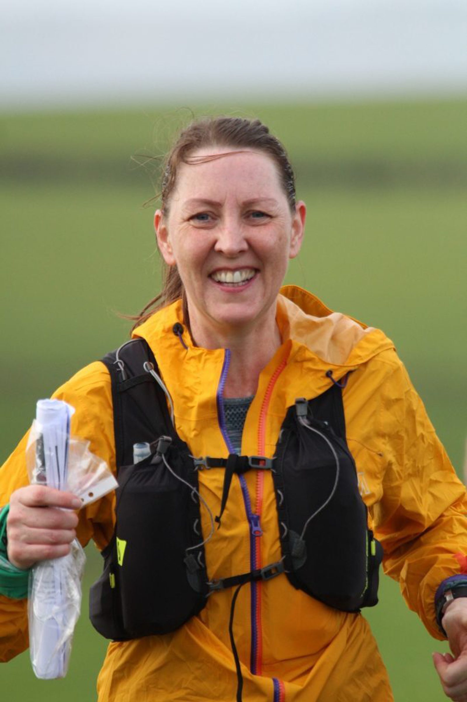 A smiling woman is wearing a bright yellow rain jacket and a black hydration vest while holding maps or papers in a plastic bag. She appears to be outdoors in a green field, likely participating in an outdoor activity or race.