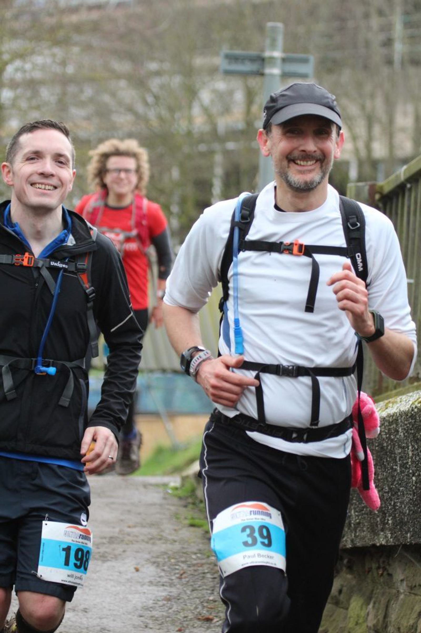 Two smiling runners, one in a black jacket and the other in a white shirt and black cap, participate in a race outdoors. Behind them, another runner in a red shirt and a cross-shaped sign are visible. The foreground shows a stone wall along the path.