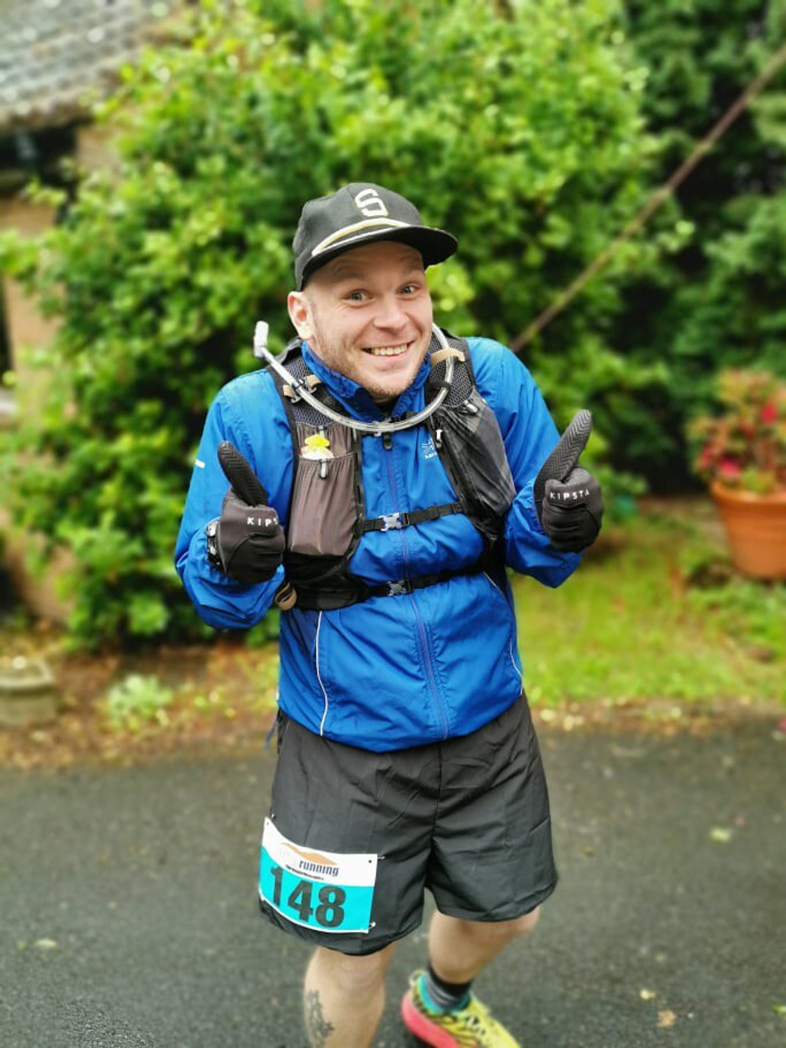 A person wearing a blue jacket, black cap, and race bib number 148 is smiling and giving two thumbs up. They are standing on a paved path with greenery and potted plants in the background. The person appears to be prepared for a race or outdoor activity.