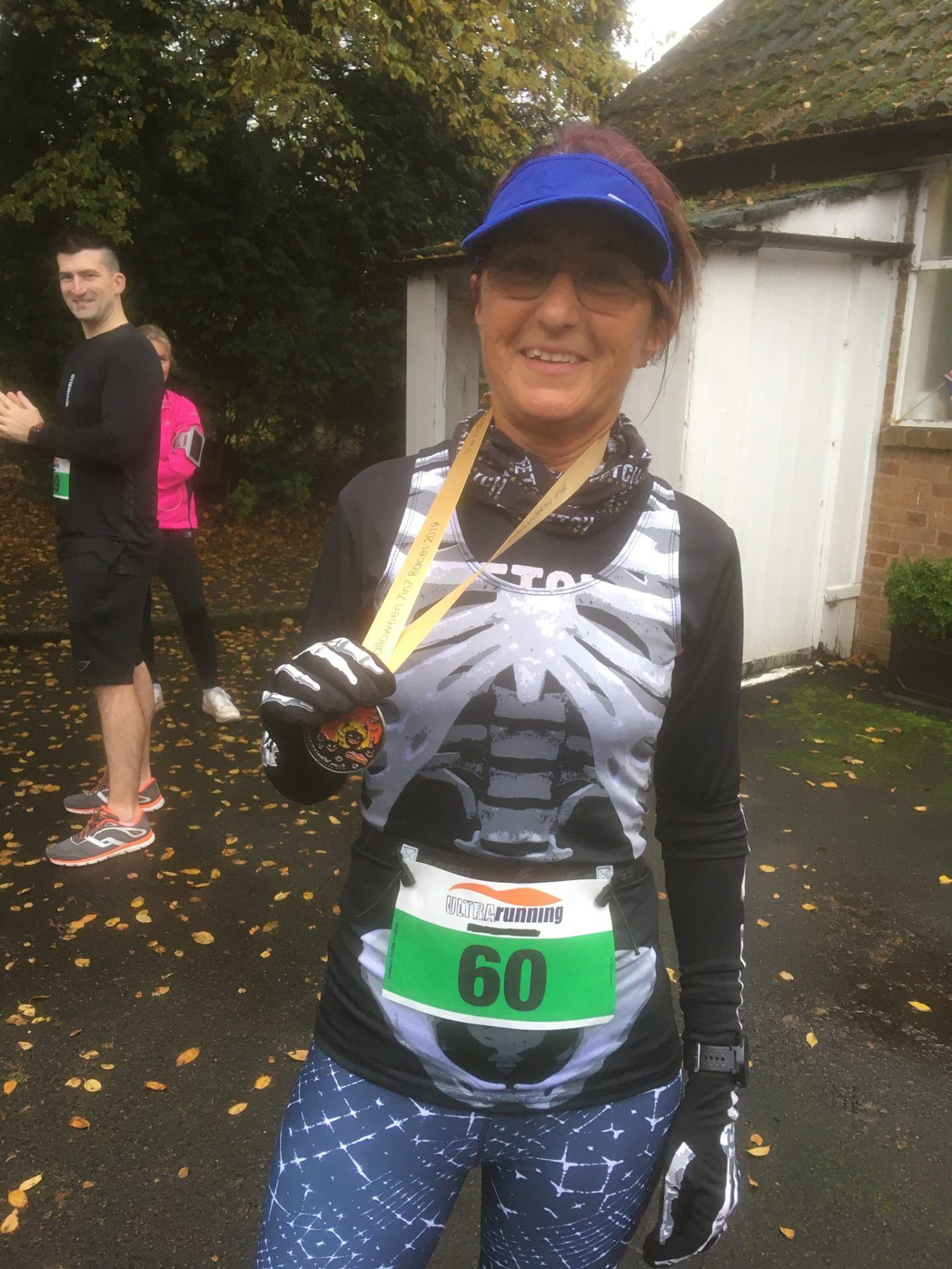 A smiling woman dressed in a skeleton-themed running shirt and blue visor holds up a medal in one hand. She wears bib number 60 and stands outdoors on a wet surface. There are other runners in the background, one wearing a pink jacket, on a rainy day.