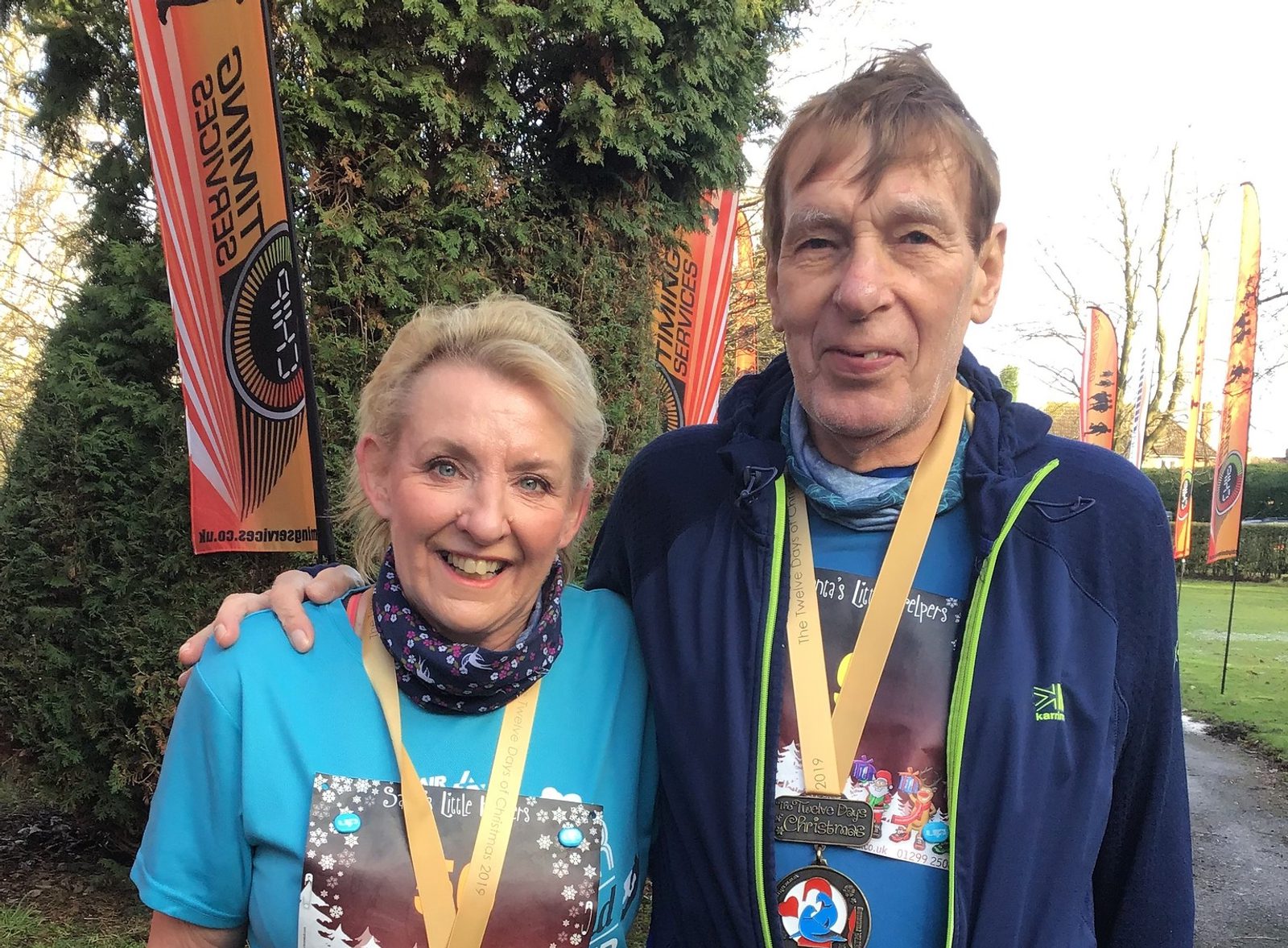 Two older adults stand outside, both smiling and wearing medals around their necks. They are dressed in athletic wear, with the woman in a blue shirt and the man in a blue jacket. Tall trees and event banners are visible in the background.