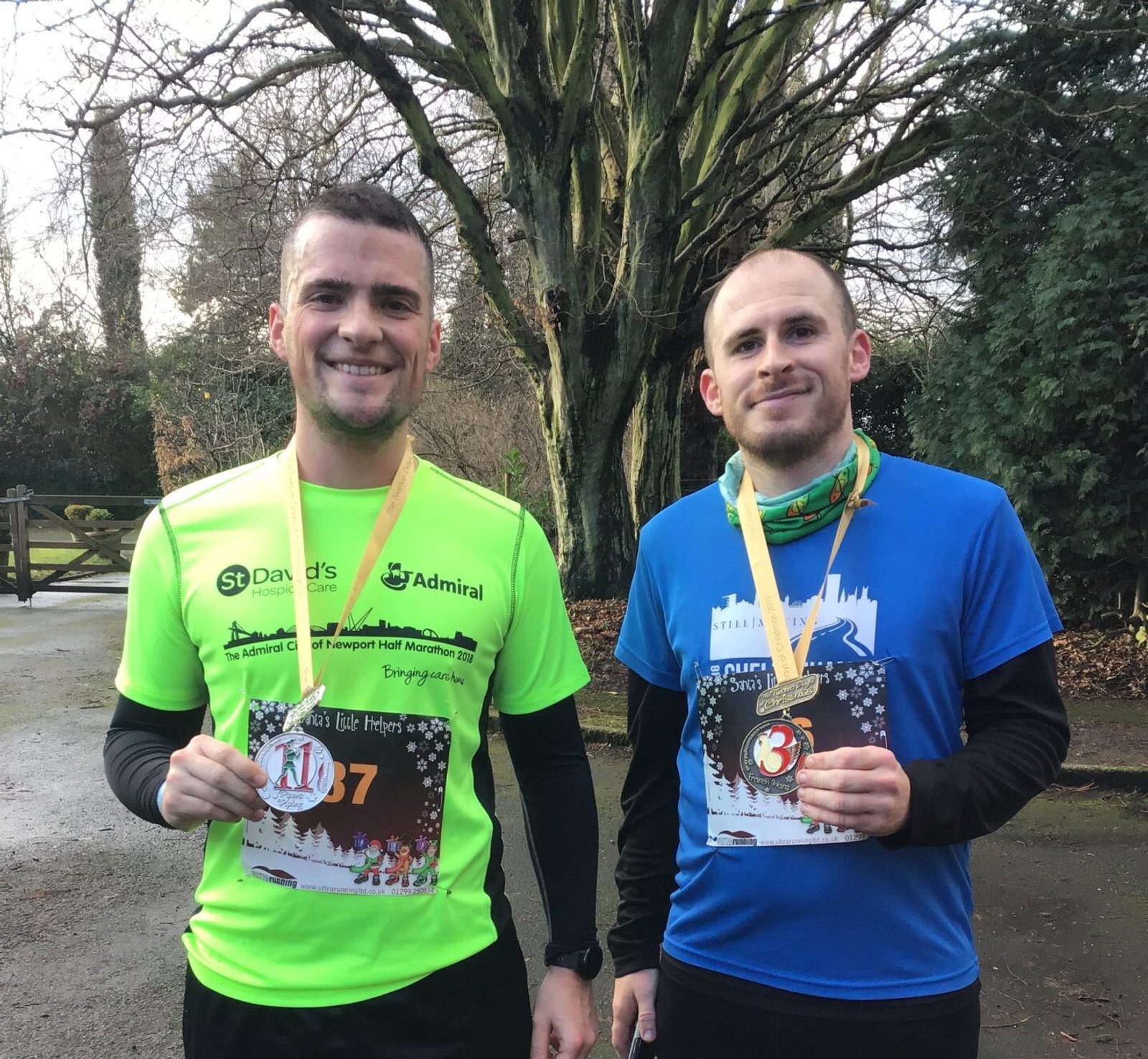 Two men are standing outside, each holding a medal and a certificate. The man on the left wears a bright green shirt with logos and the number 1427, while the man on the right wears a blue shirt. Both are smiling; trees and outdoor scenery are visible in the background.