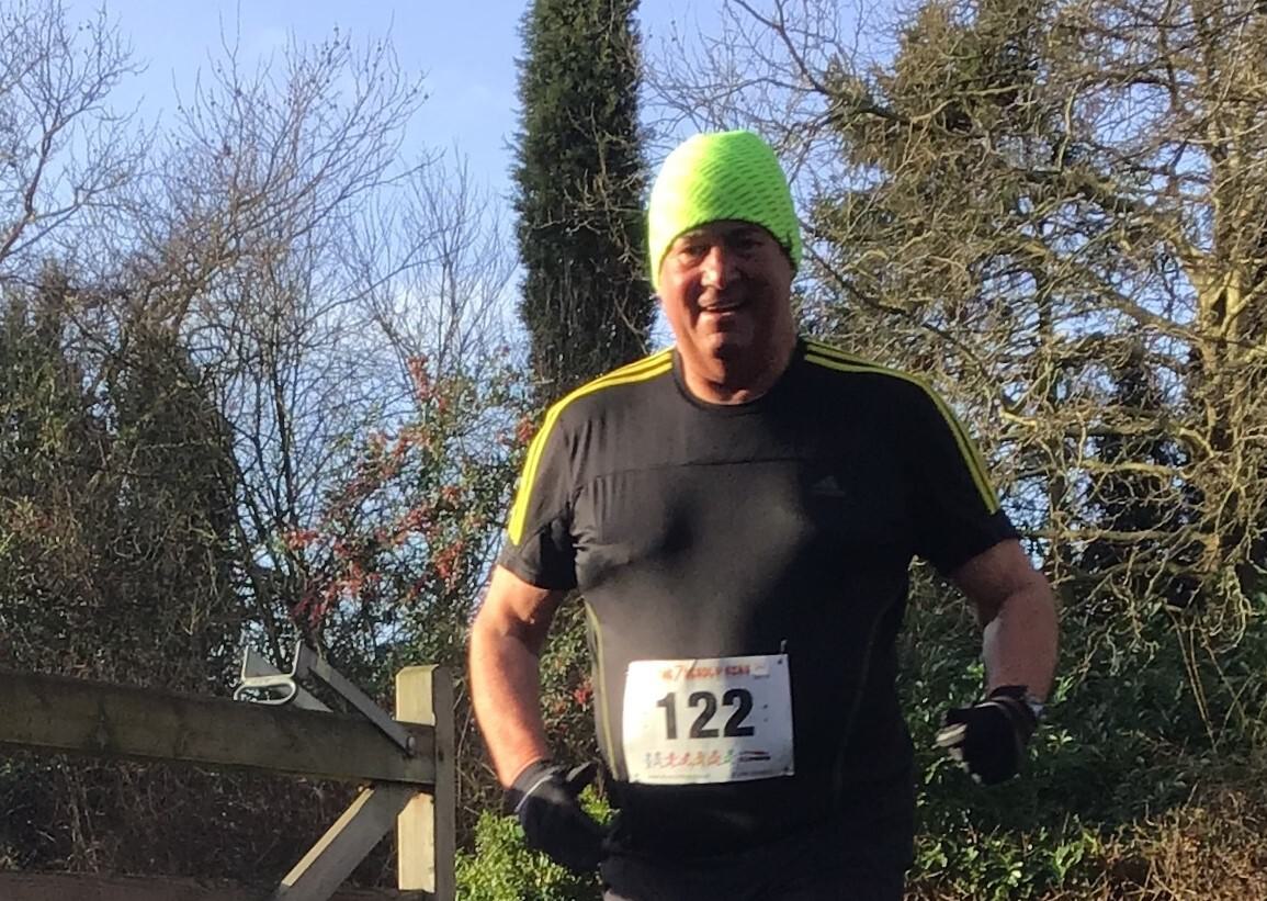 A man wearing a neon green beanie, a black shirt with yellow stripes, and a race bib with the number 122 is jogging outdoors. Trees and a wooden fence are visible in the background on a clear day.