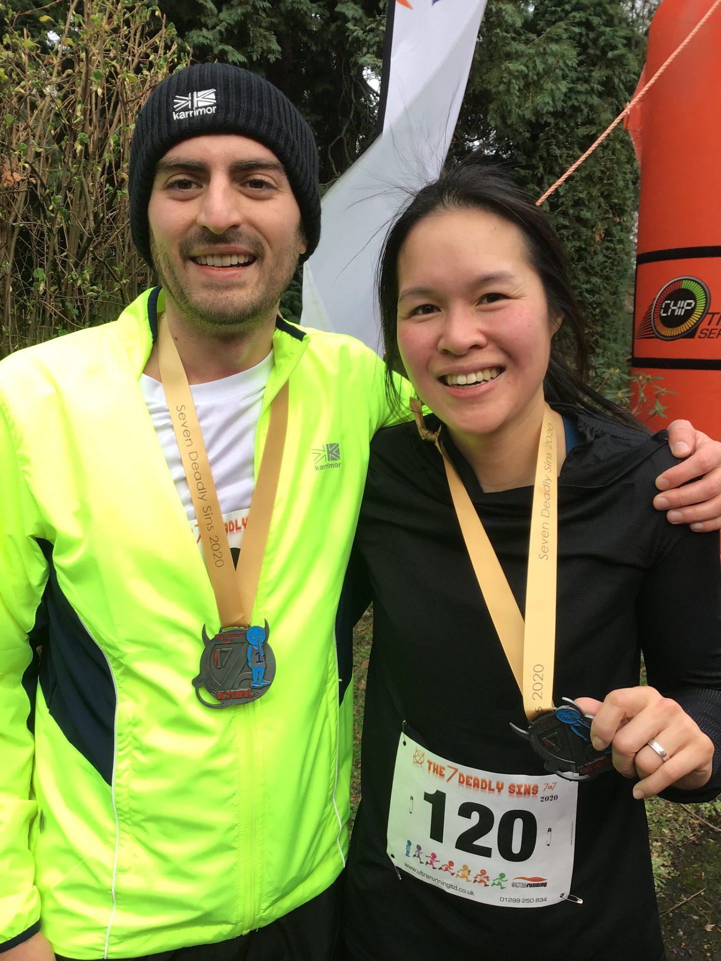 Two smiling marathon runners pose for a photo with their medals. The man, wearing a neon yellow jacket and black beanie, stands beside a woman in a black top and race bib number 120. They are outdoors, near an inflatable race finish arch.