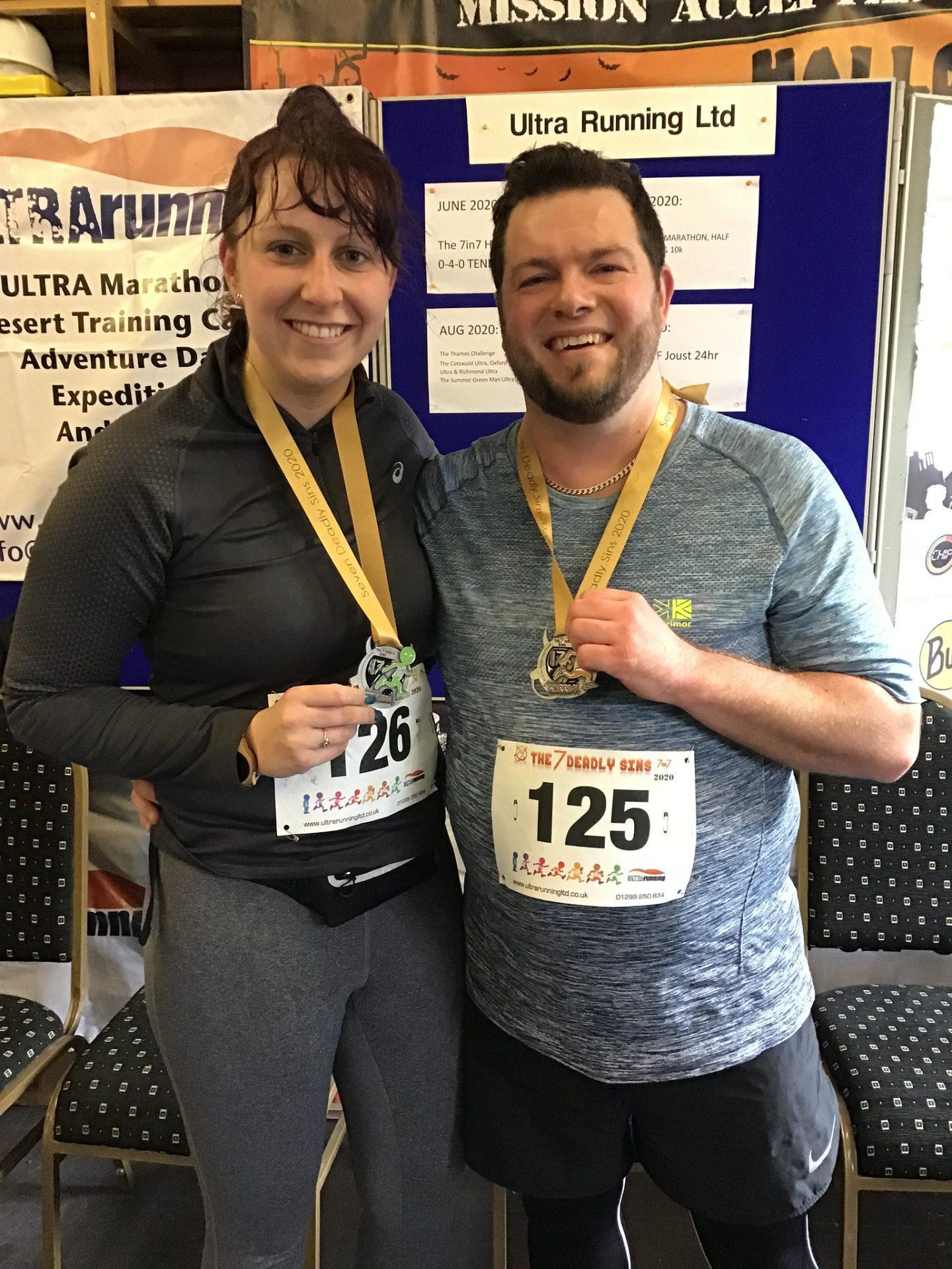 Two smiling individuals are standing close together, each holding a medal. Both are wearing race bibs, the woman with the number 126 and the man with the number 125. They are indoors, with an event board for Ultra Running Ltd in the background.