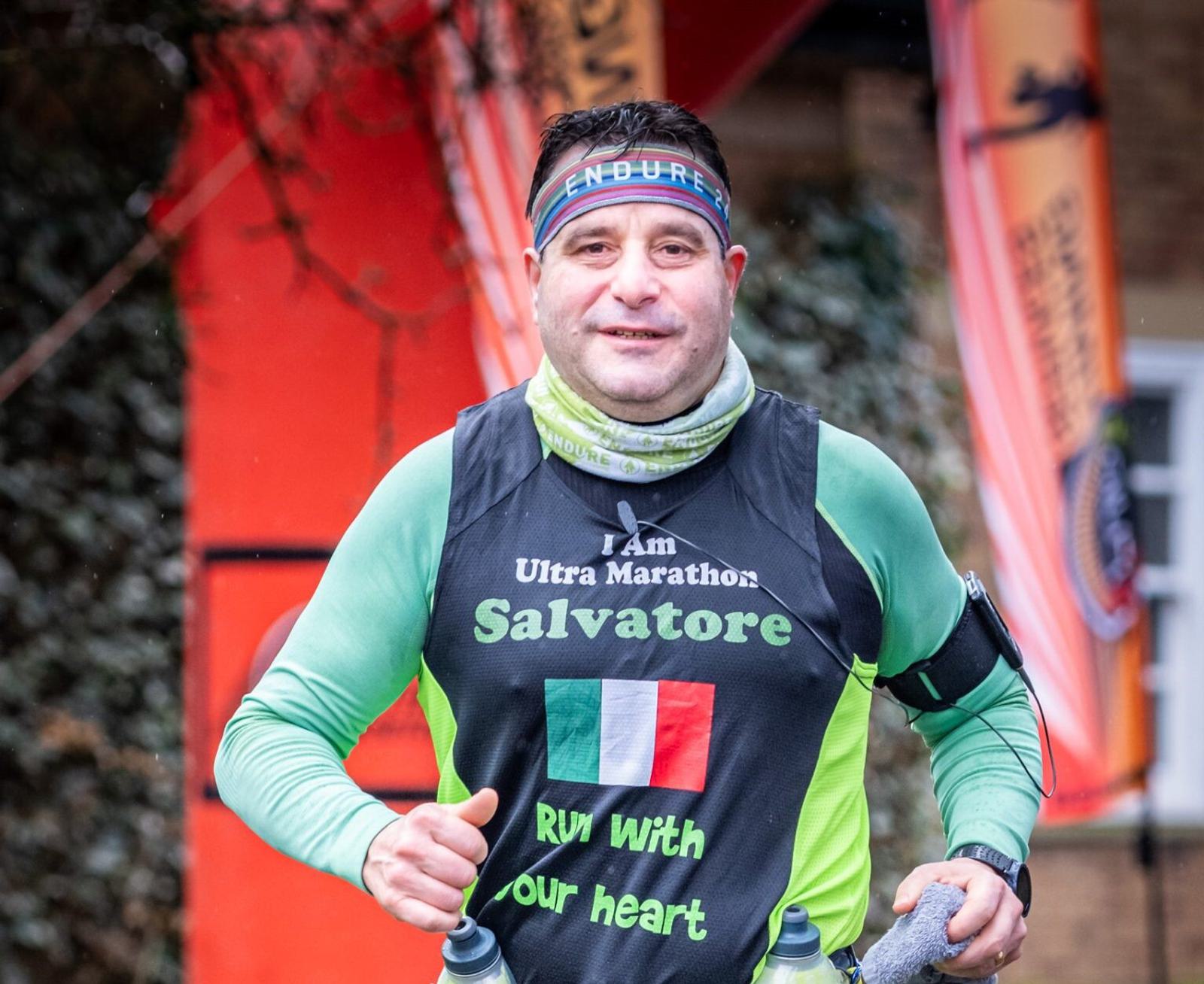 A runner wearing a black and green outfit bearing the words "I am Ultra Marathon Salvatore" and an Italian flag on the front is running. They are carrying two bottles in their vest. An orange banner and greenery are visible in the background.