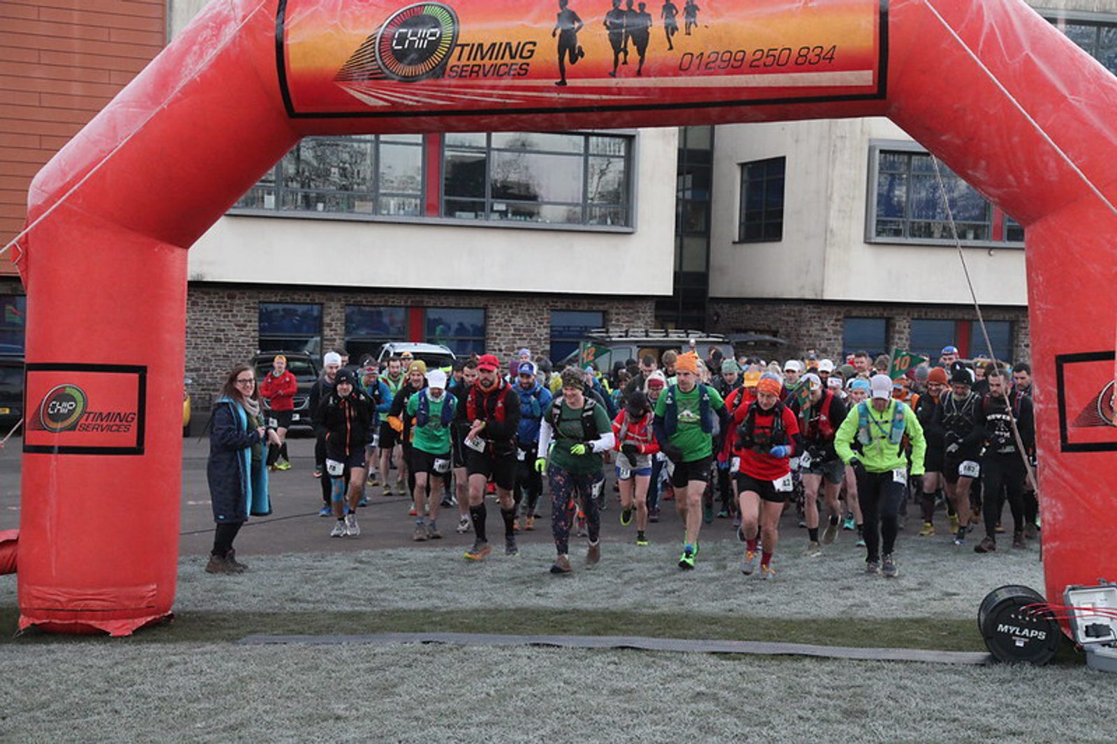 A group of runners bundled in cold-weather gear starts a race beneath a large red inflatable arch with a timing services banner, outside a modern building on a frosty morning.