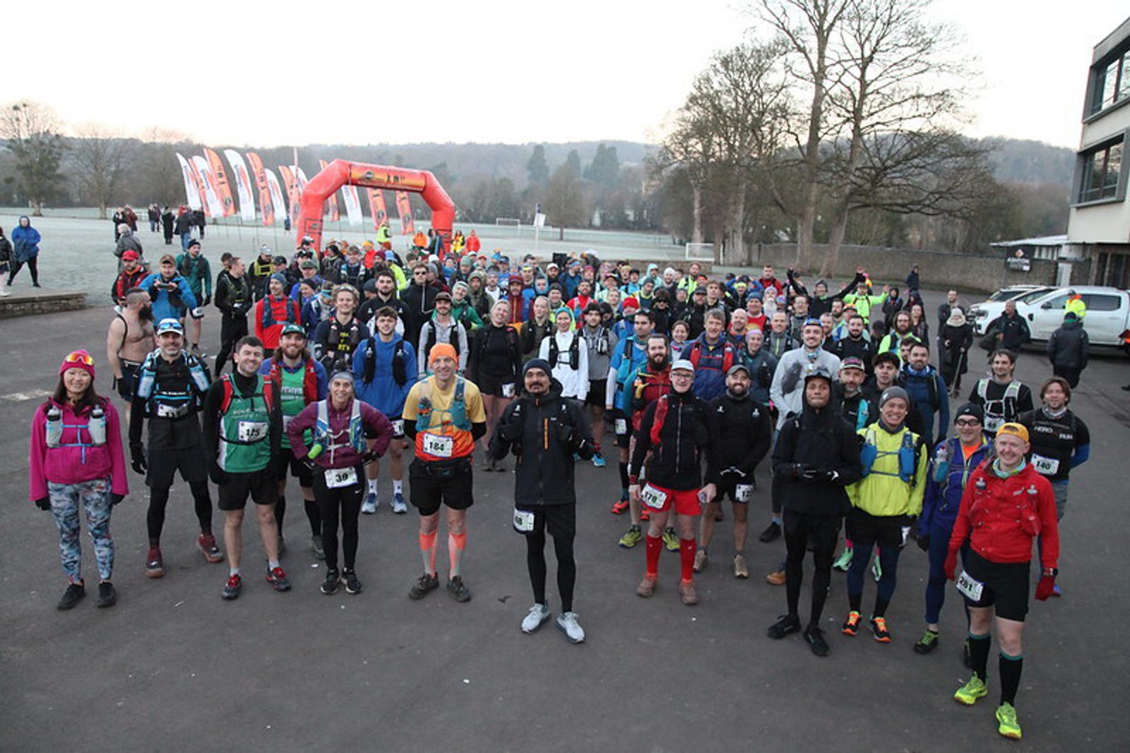 A large group of runners wearing race bibs and athletic gear stand together outdoors, smiling and posing before a race. An orange inflatable arch and flags are visible in the background on a clear day.
