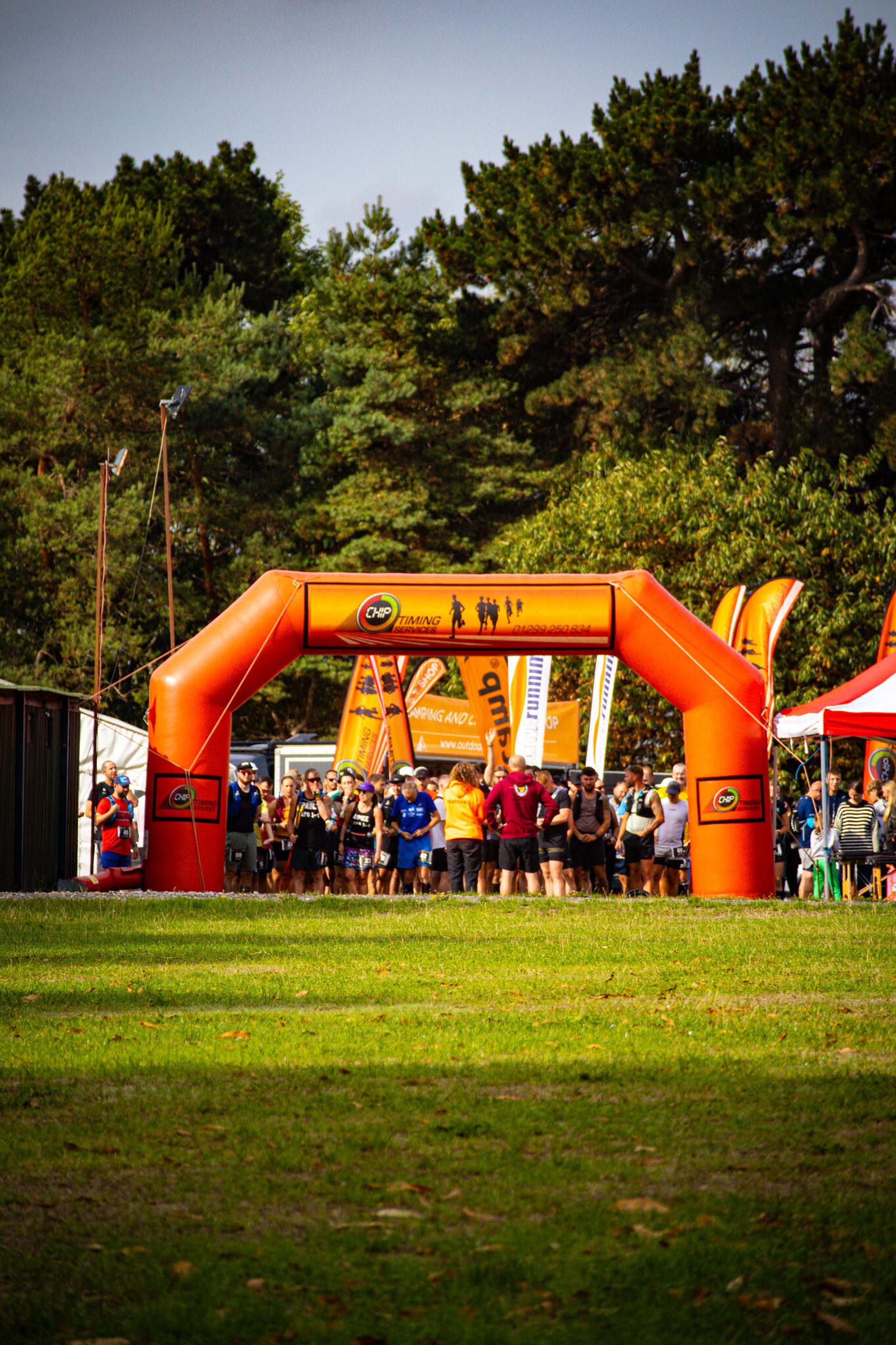 A group of runners gathers at the starting line under an orange inflatable archway at an outdoor race event, surrounded by trees and green grass.