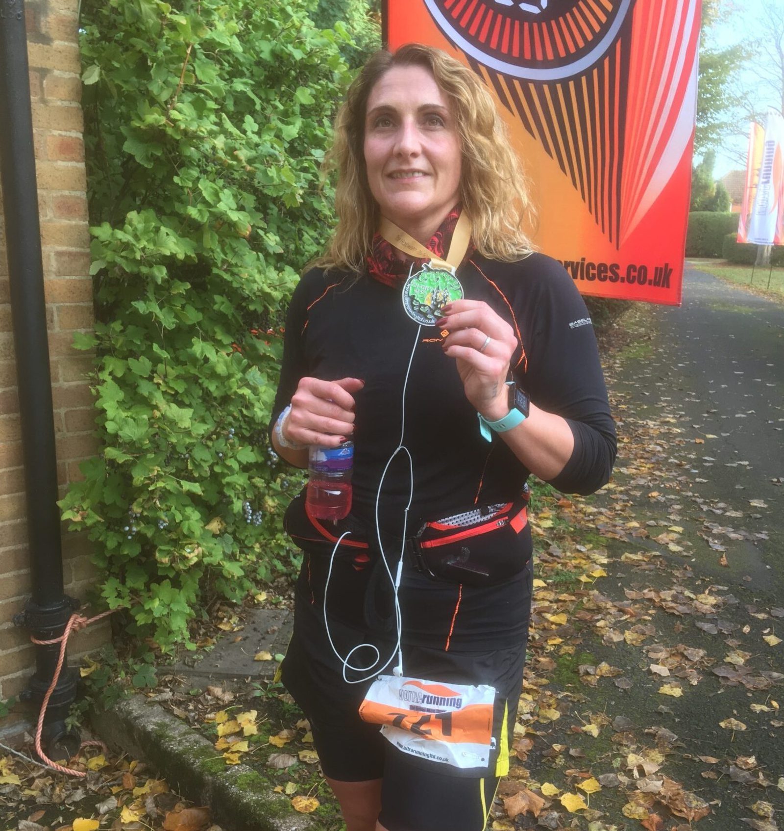 A woman with wavy blonde hair stands outdoors, holding a green medal and a water bottle. She's wearing a black outfit with red accents and has earphones connected to a device. There's a colorful banner in the background and some fallen leaves on the ground.