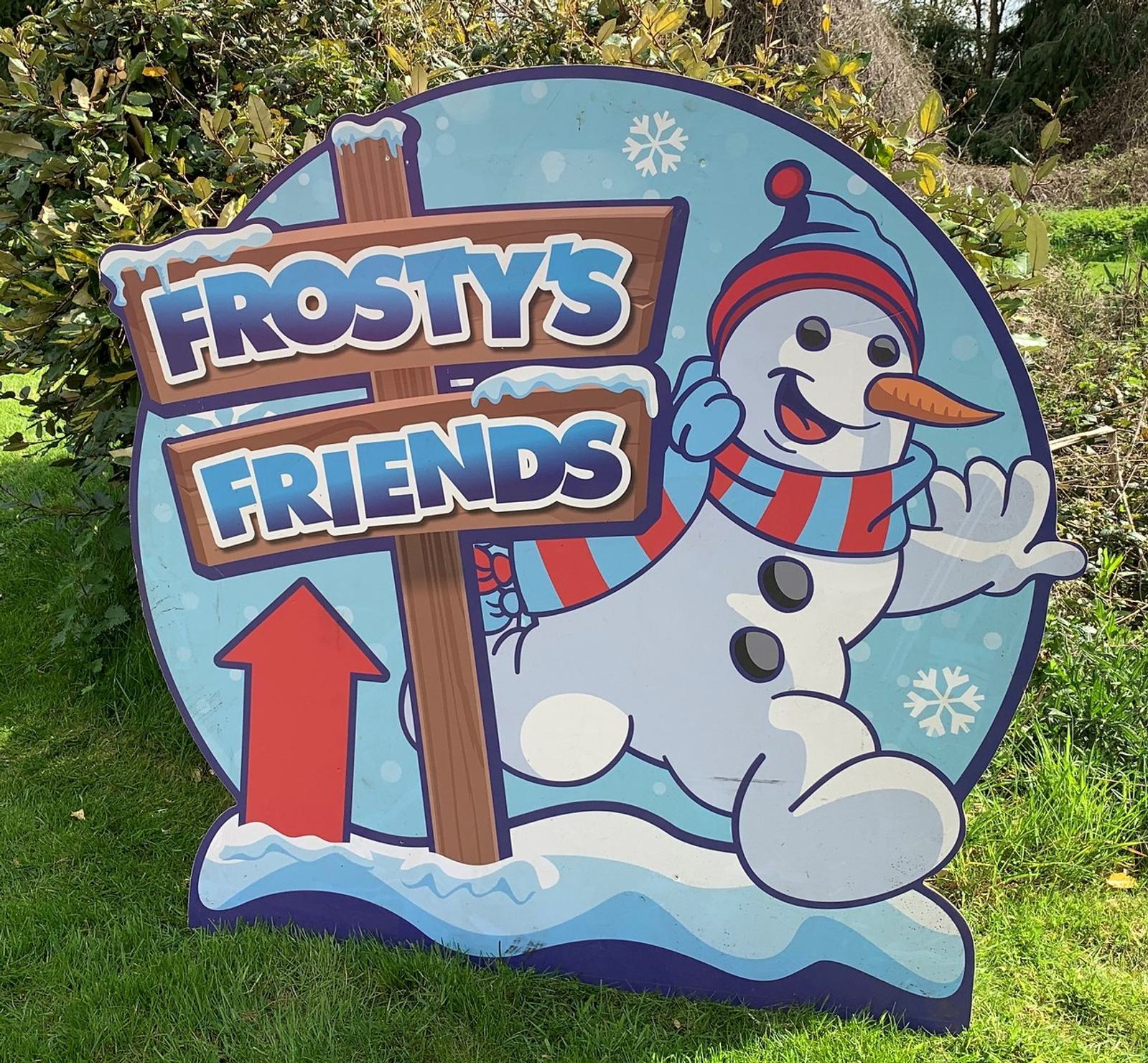 A colorful sign in a grassy area features a smiling snowman with a carrot nose, black buttons, a striped red and blue scarf, and a matching hat. The sign reads "Frosty's Friends" with a red arrow pointing up, all set against a winter-themed background.