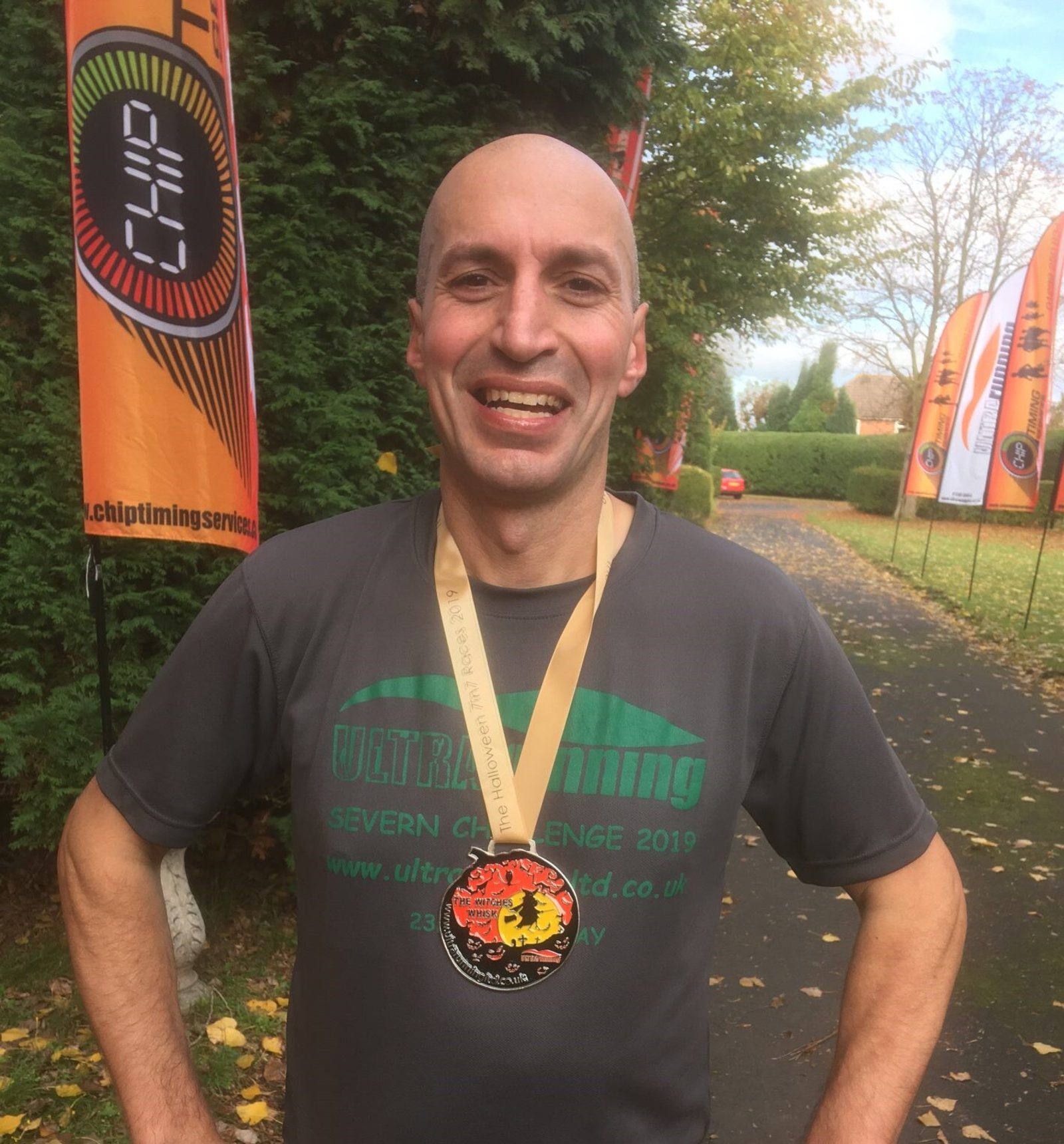 A smiling man stands outdoors, wearing a gray "Severn Challenge 2019" T-shirt and a medal around his neck. Orange and black flags line the path in the background, with green trees and scattered autumn leaves on the ground.