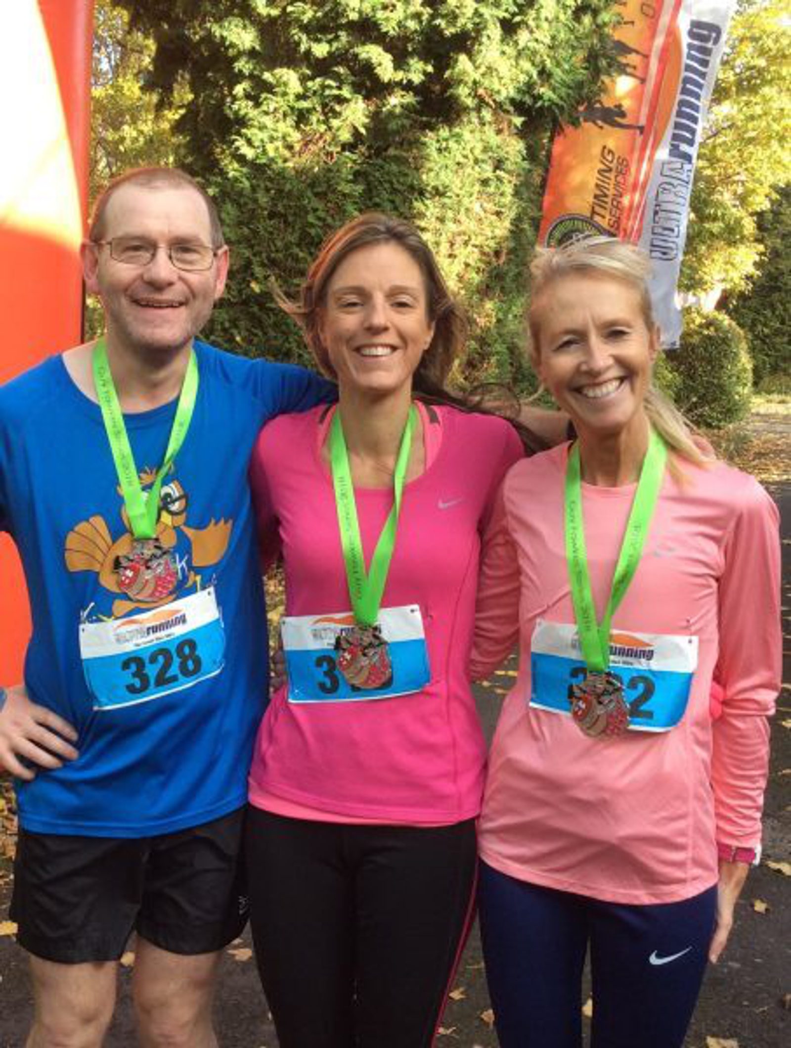 Three runners smile for the camera after a race, each wearing a medal and bib numbers 328, 3, and 232. They stand together, with one man in a blue shirt (left) and two women wearing pink shirts (center and right), against a backdrop of trees and race banners.